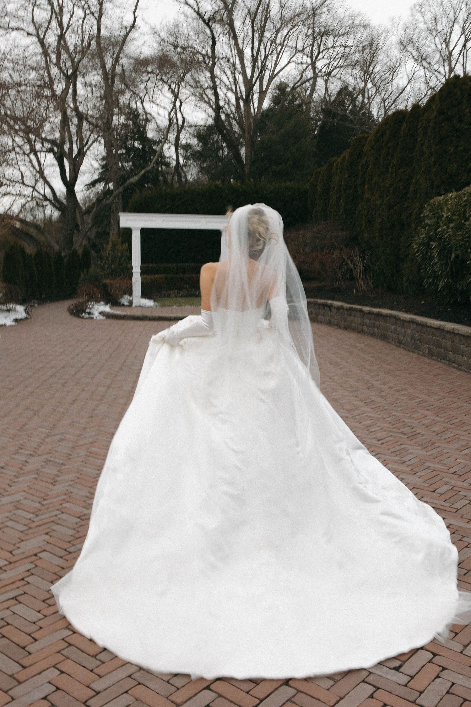 A bride in a flowing white wedding gown and veil stands on a brick pathway near a white arbor.
