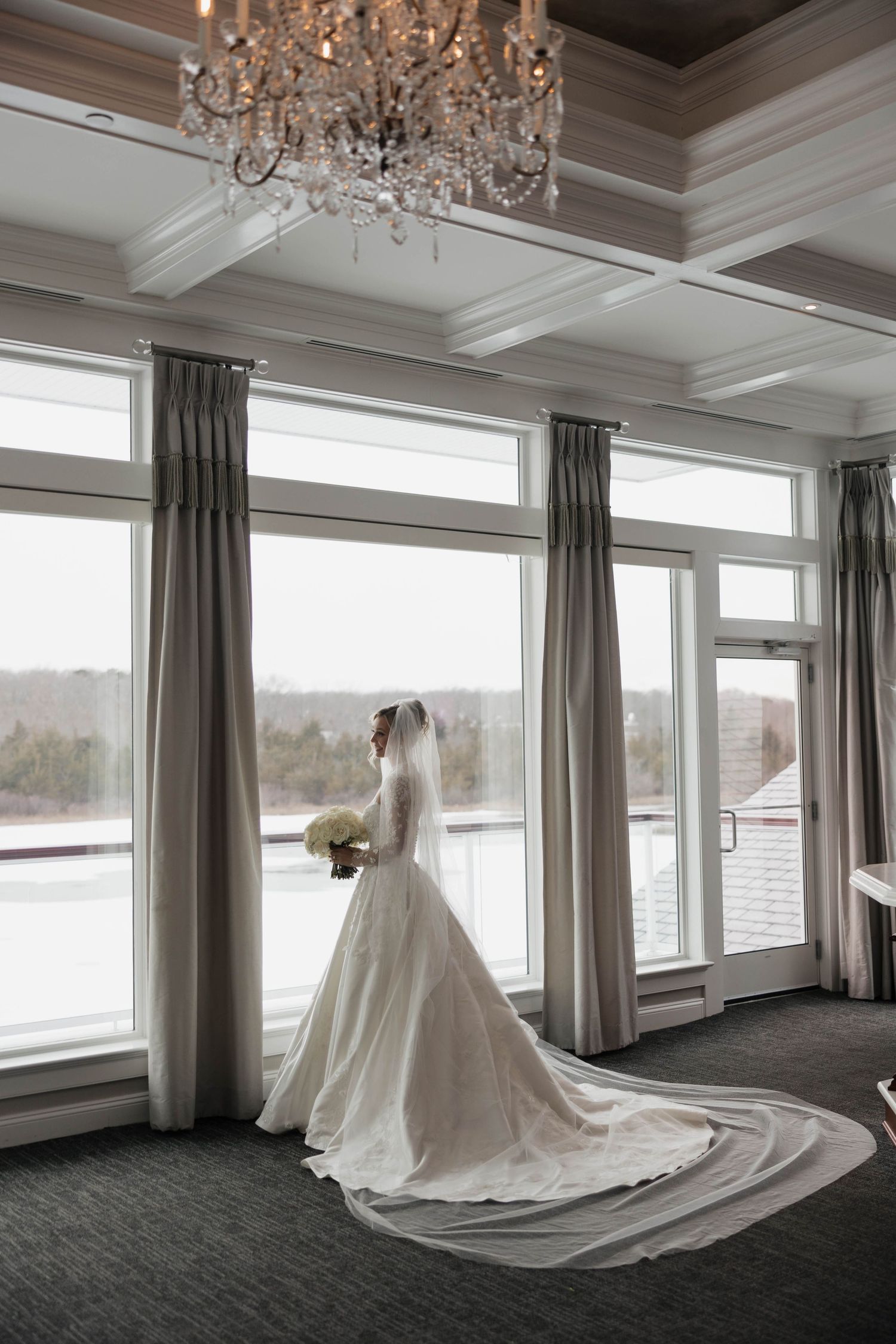 Dramatic full-length wedding dress silhouette in luxurious room with chandelier and water view.