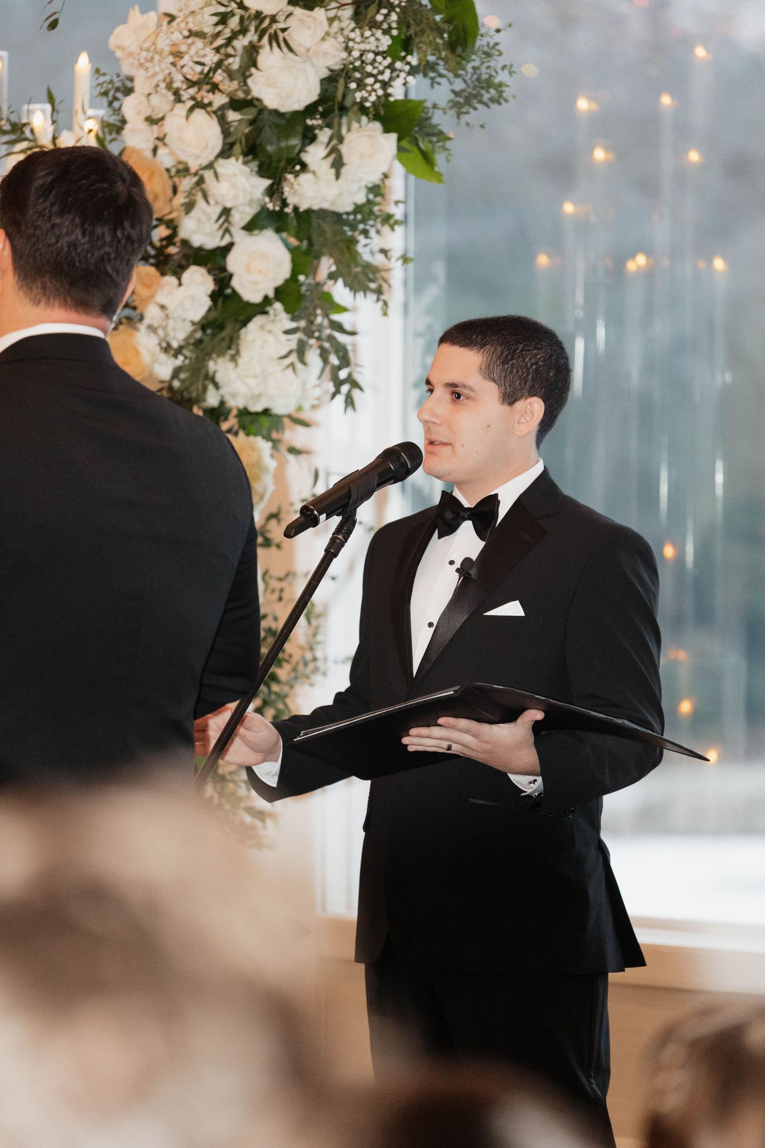 Speaker in black suit gestures while delivering speech at formal wedding ceremony.