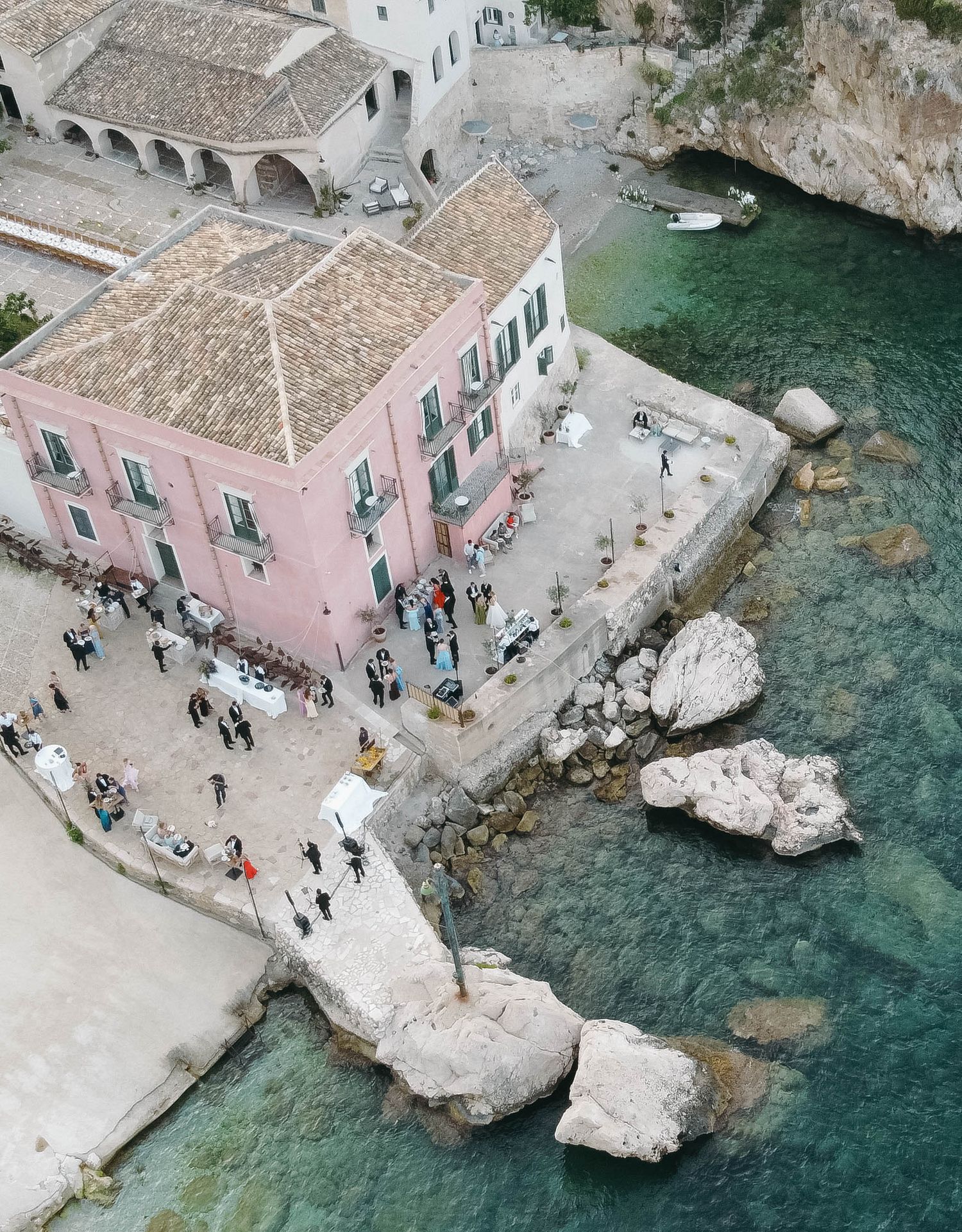 Mediterranean coastal scene with pink buildings, stone paths and visitors near crystal waters.