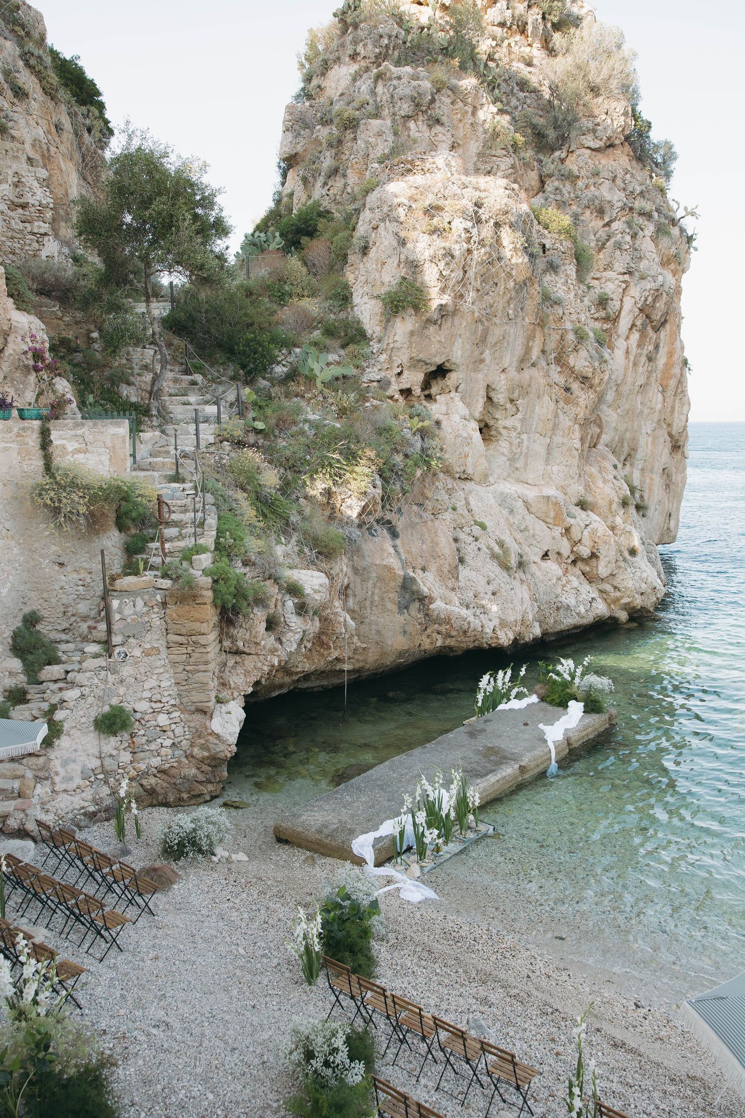 Intimate wedding ceremony setup on a secluded rocky beach cove with wooden chairs and white floral decorations.