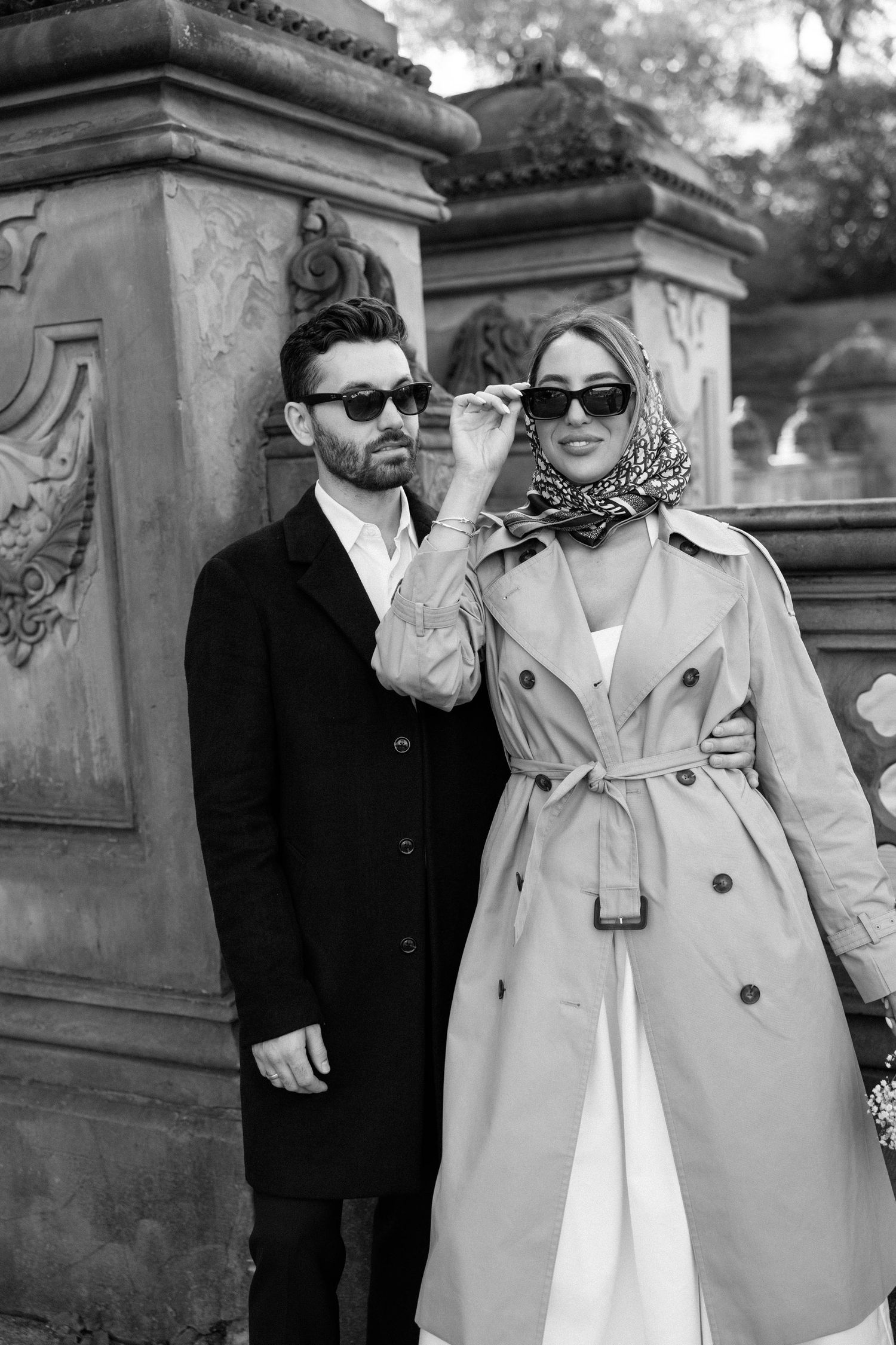 A couple wearing sunglasses and light colored coats poses together outdoors.