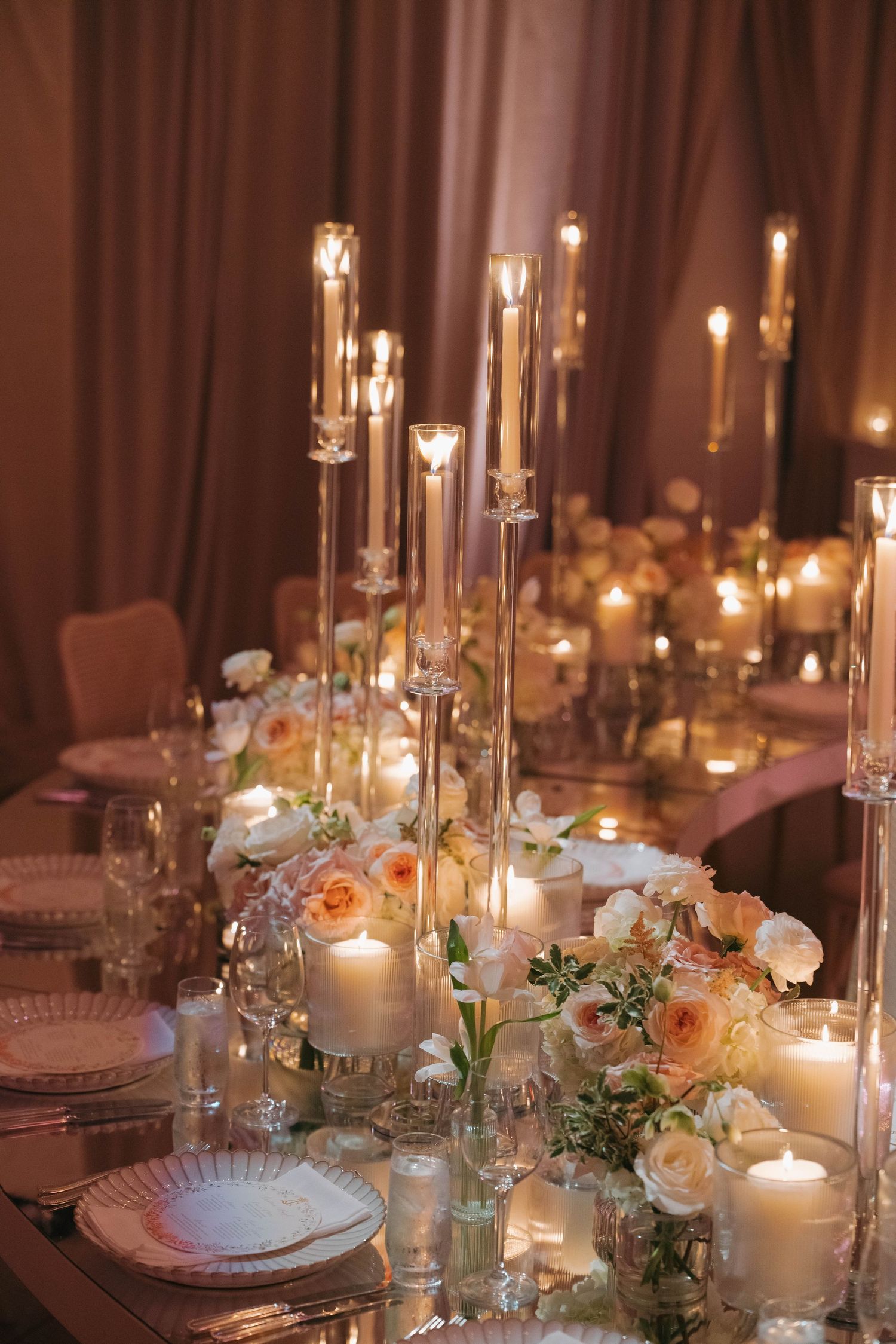 Close-up of wedding table decor featuring glass candleholders, white candles, and coral roses.