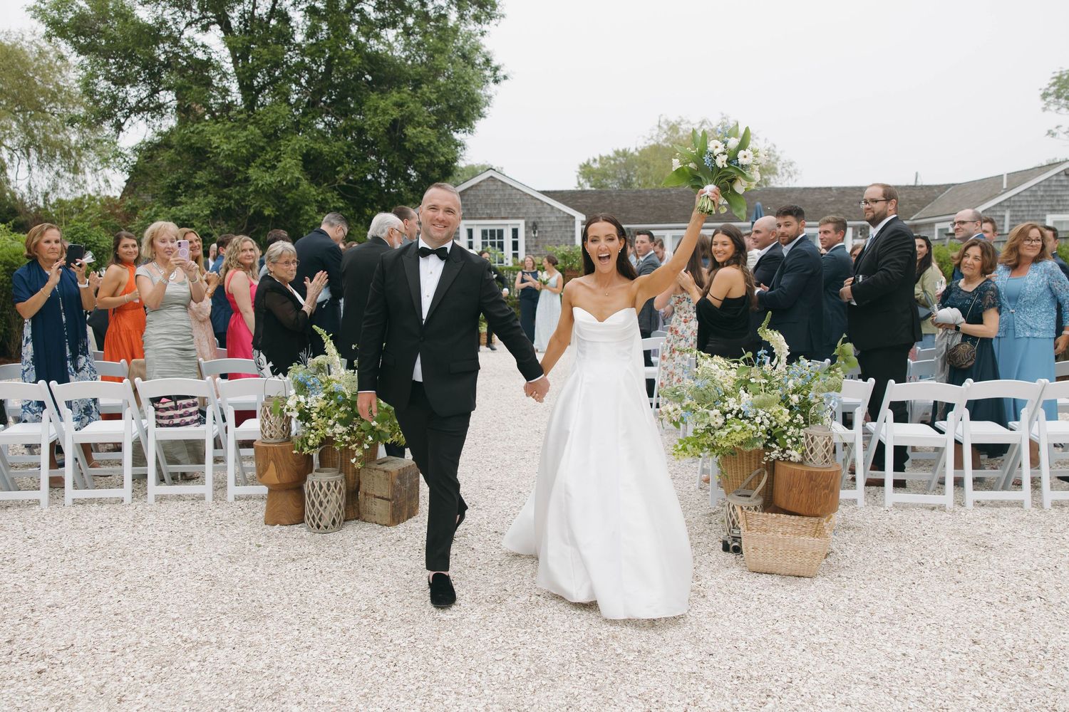 Panoramic view of wedding ceremony with guests seated on white chairs on gravel ground.