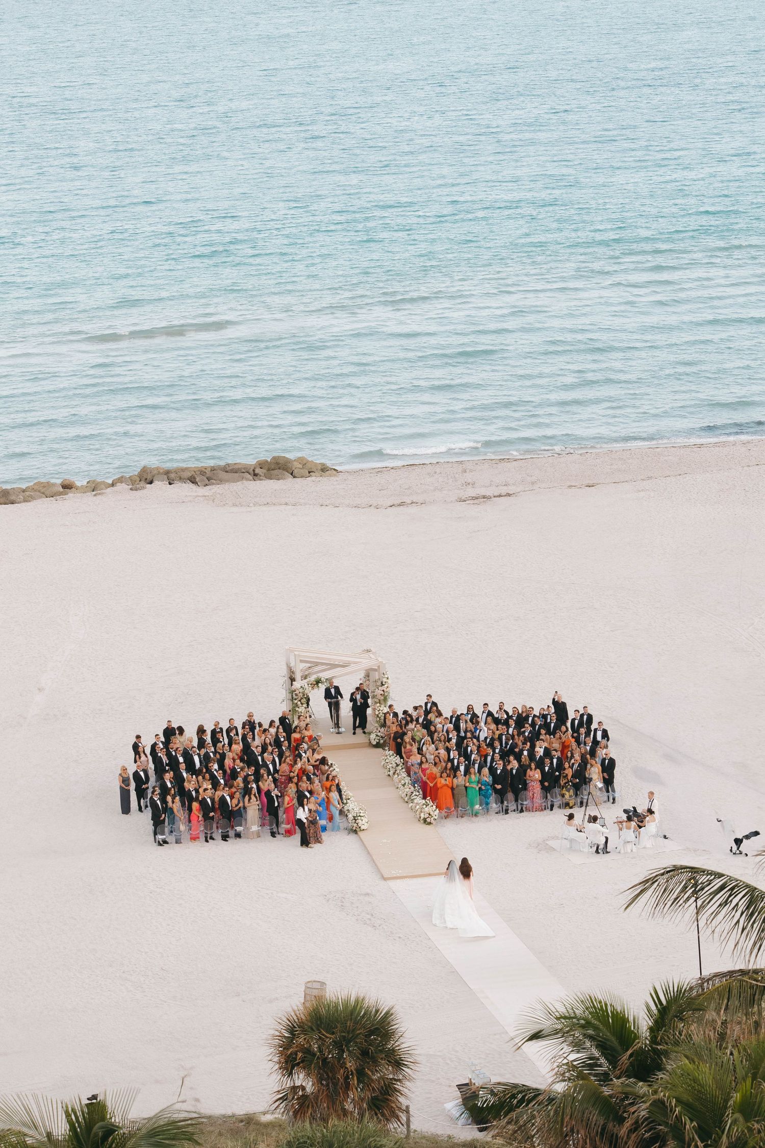 Aerial view of wedding guests gathered on a pristine beach forming organized group formations.