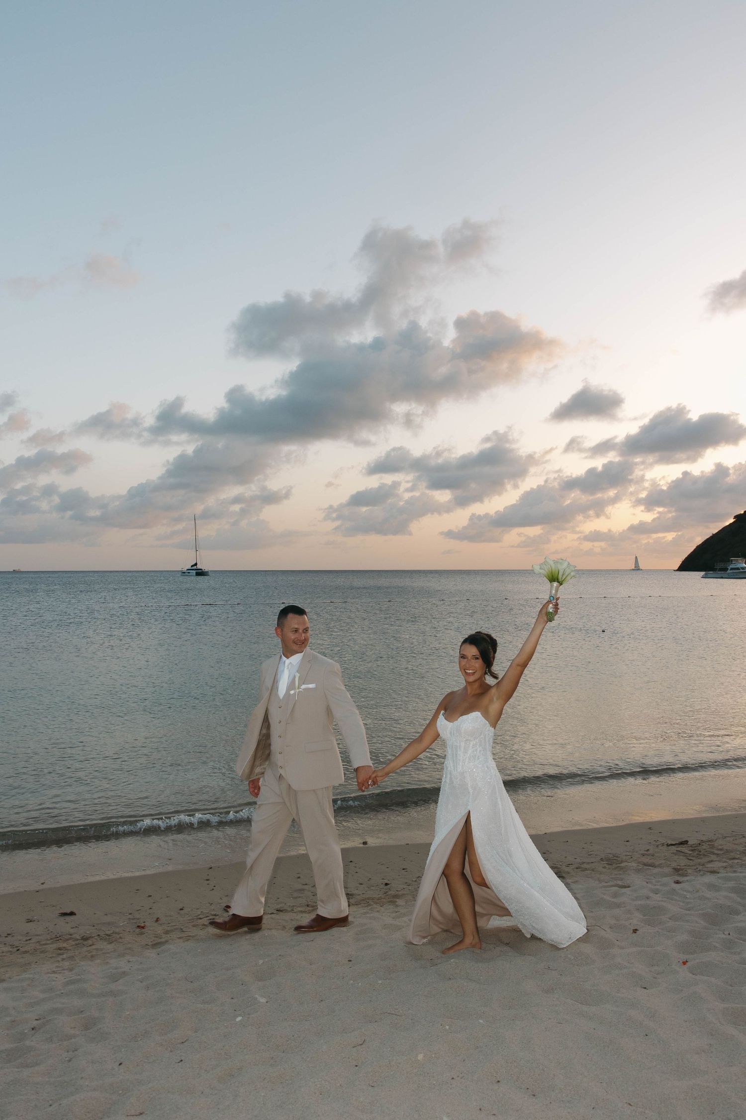 Bride and groom walk hand-in-hand on a sandy beach at sunset, bride raising bouquet in celebration.