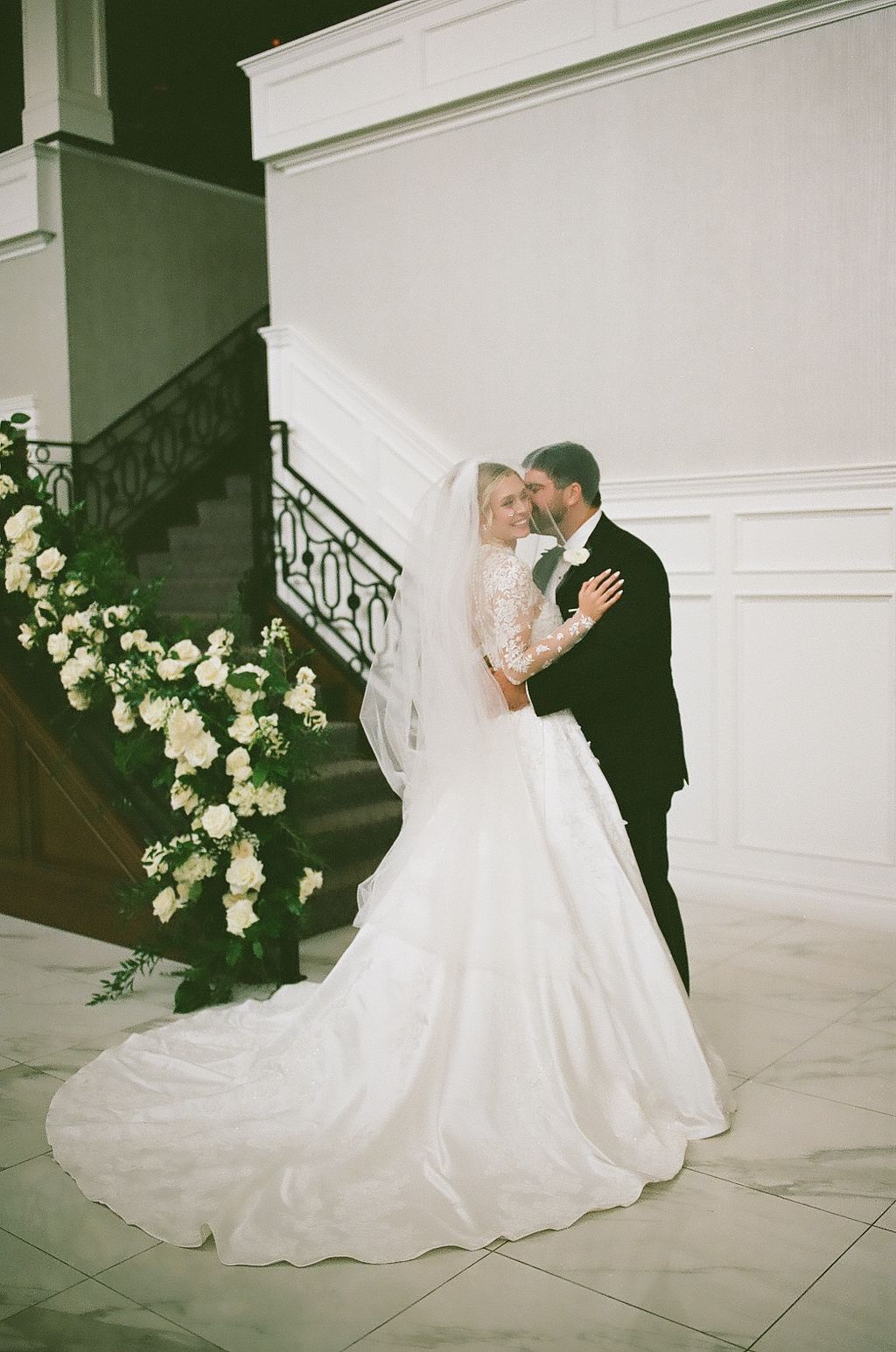 A bride and groom share an intimate moment in a series of romantic wedding photos against a white wall with floral decor.