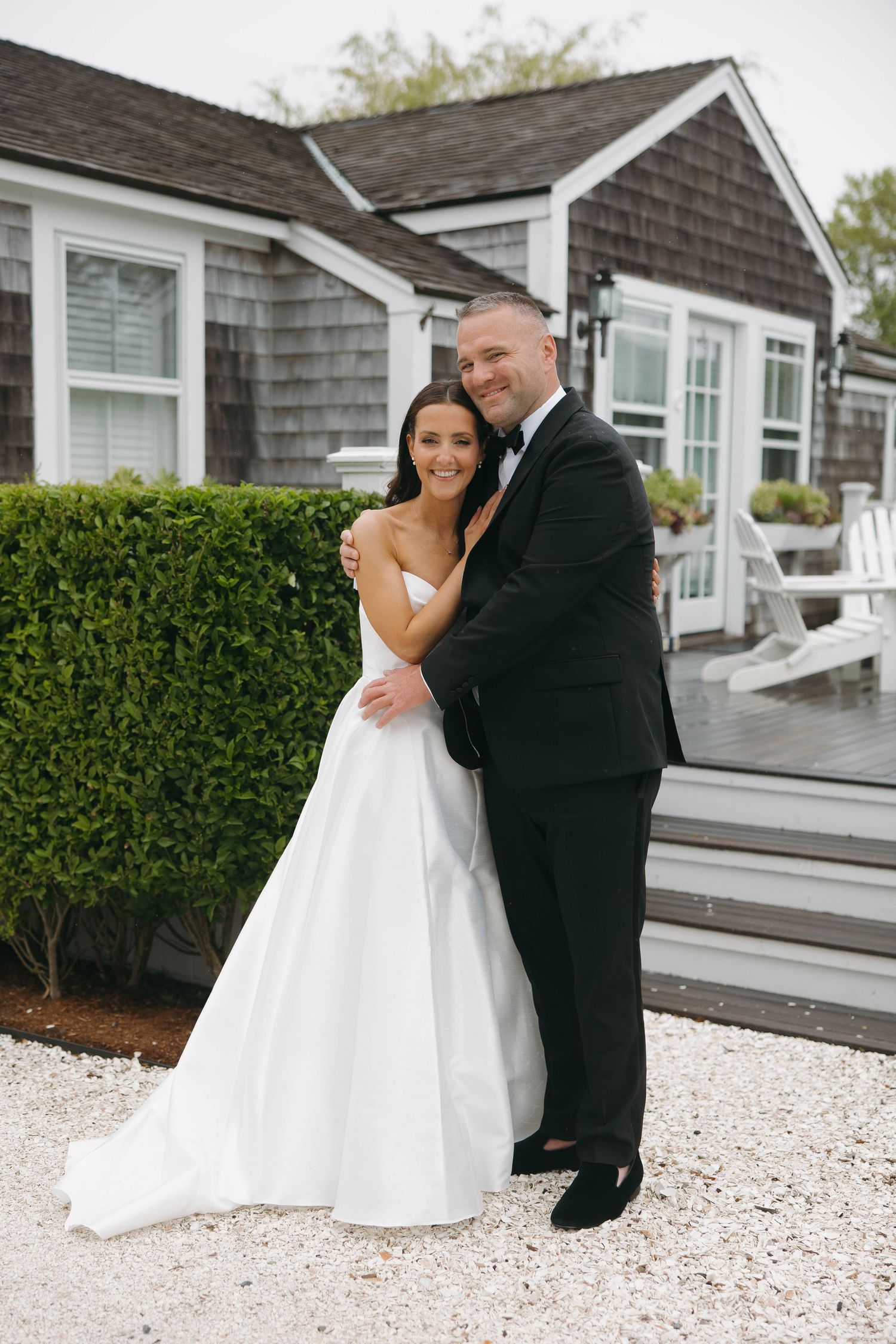 Newlyweds embrace on white gravel path outside classic New England shingle-style building.
