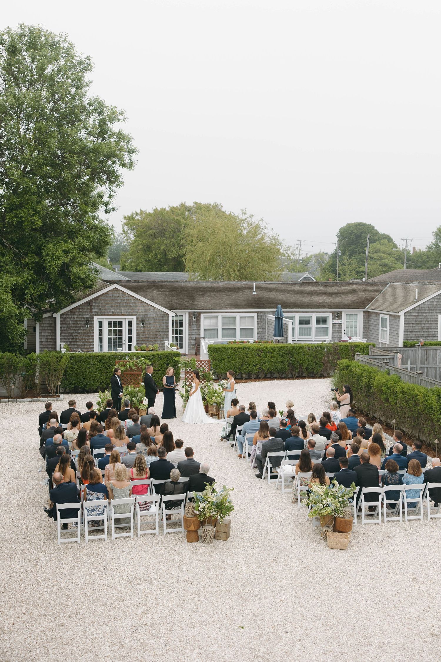 Guests seated at outdoor wedding ceremony with white chairs and coastal style buildings.