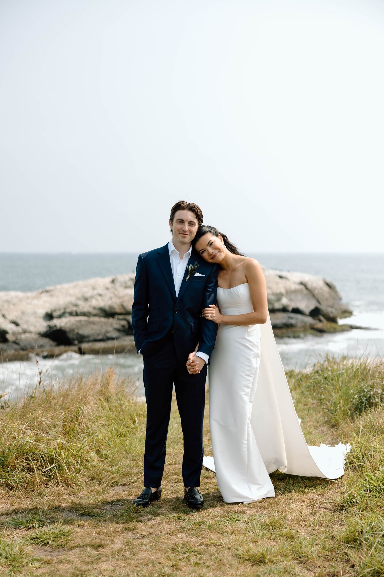 Couple in wedding attire standing together on rocky coastal cliff overlooking ocean.