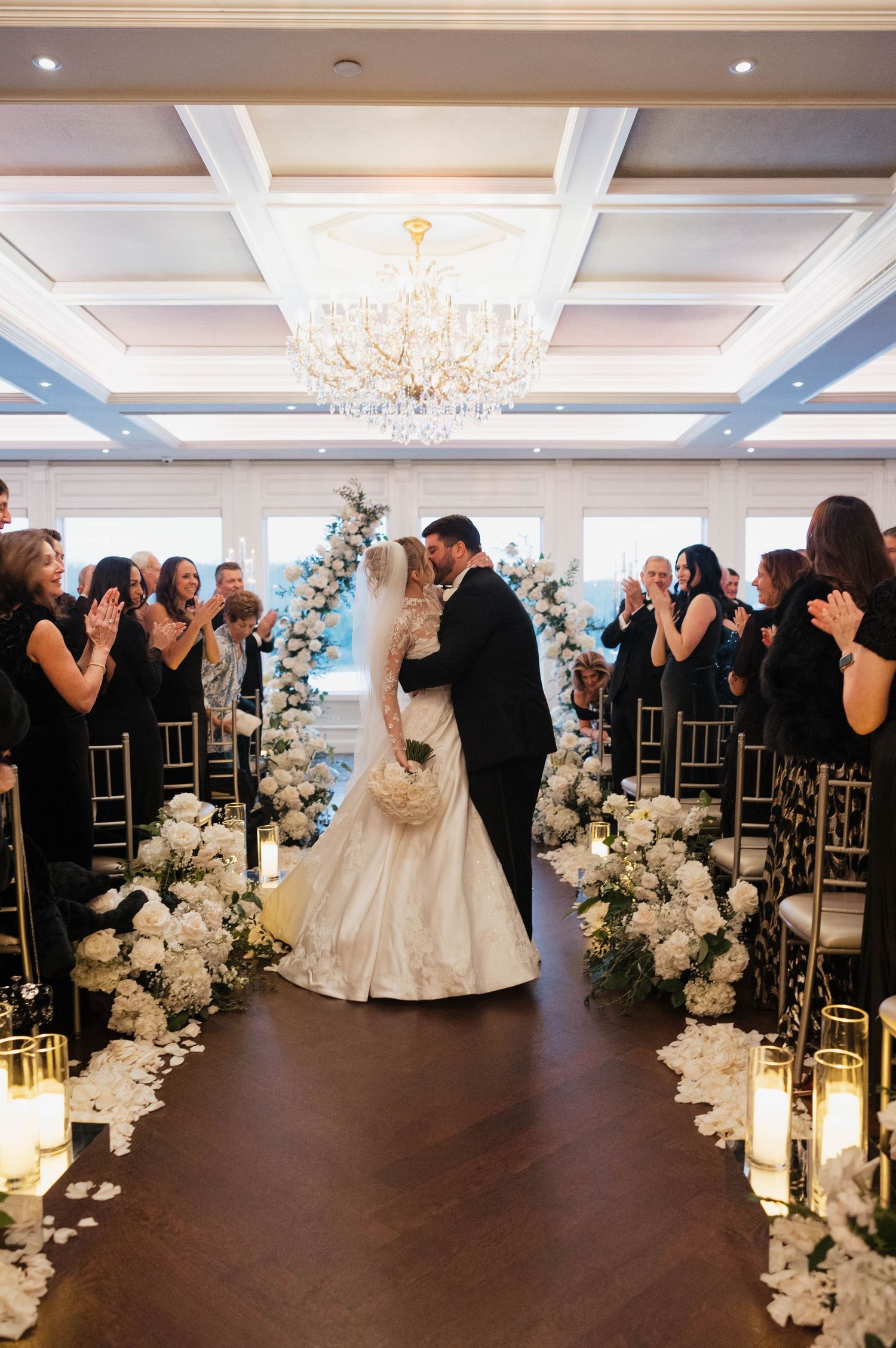 Wedding guests applaud as newlyweds share a moment under crystal chandelier.
