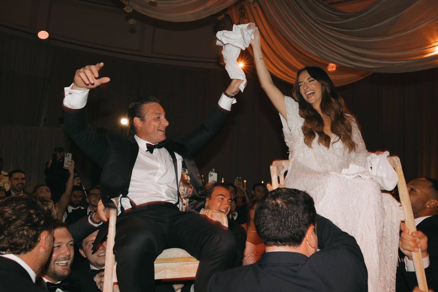 Wedding celebration with guests lifting celebrants on chairs during traditional dance.