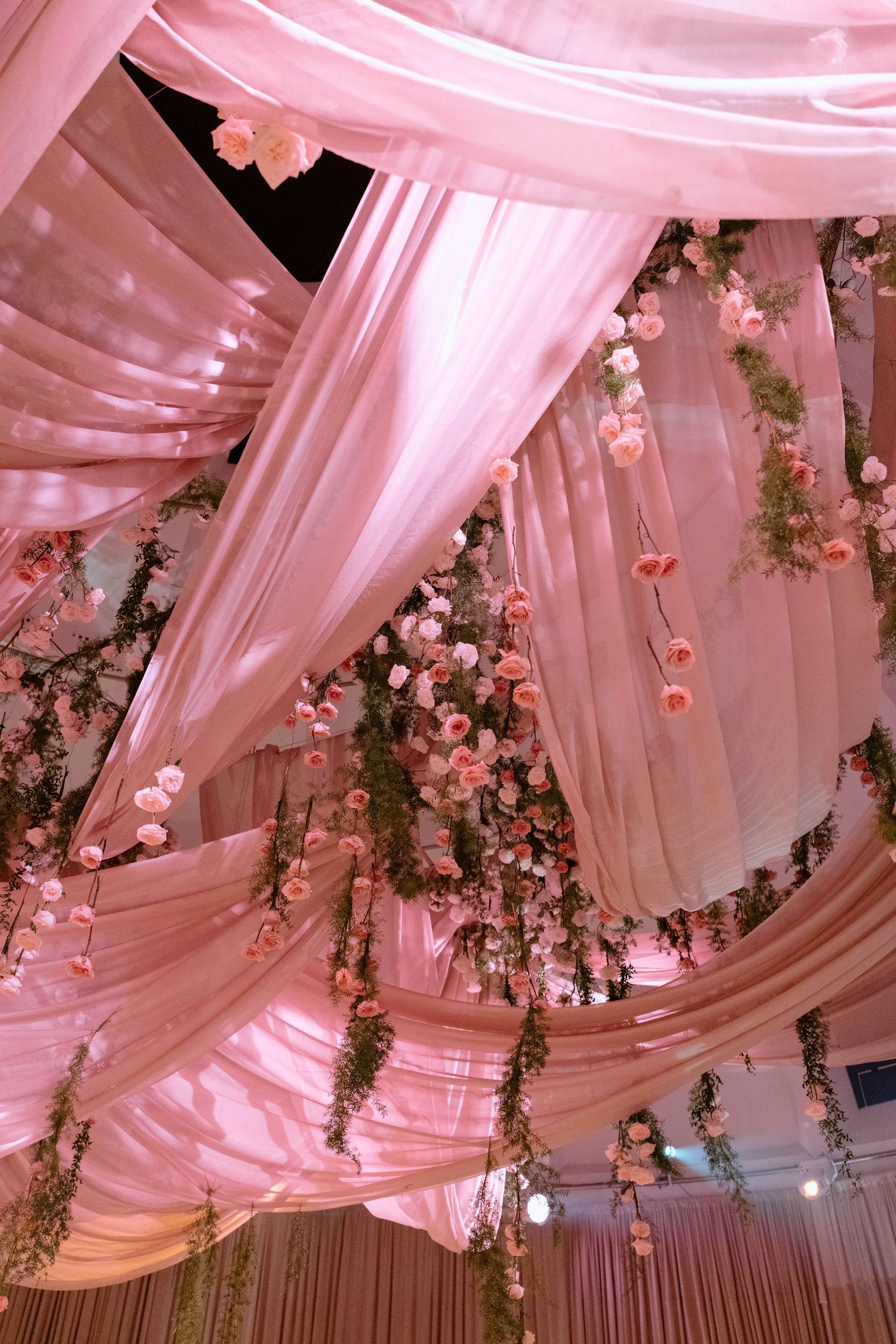 Ethereal ceiling draping with cascading floral installations and pink uplighting creating a romantic atmosphere.