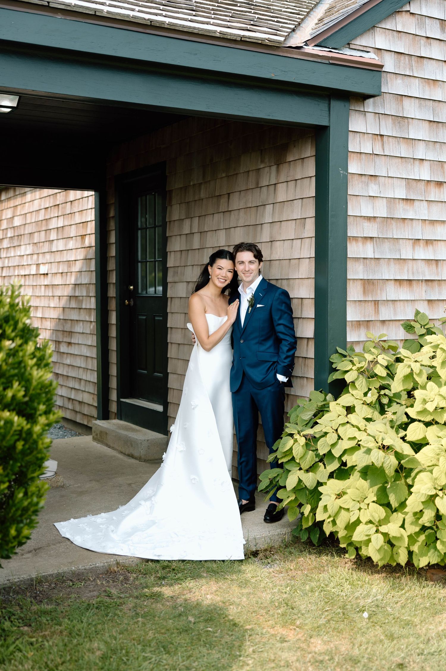 A wedding couple poses against a shingled building with green trim and yellow foliage.
