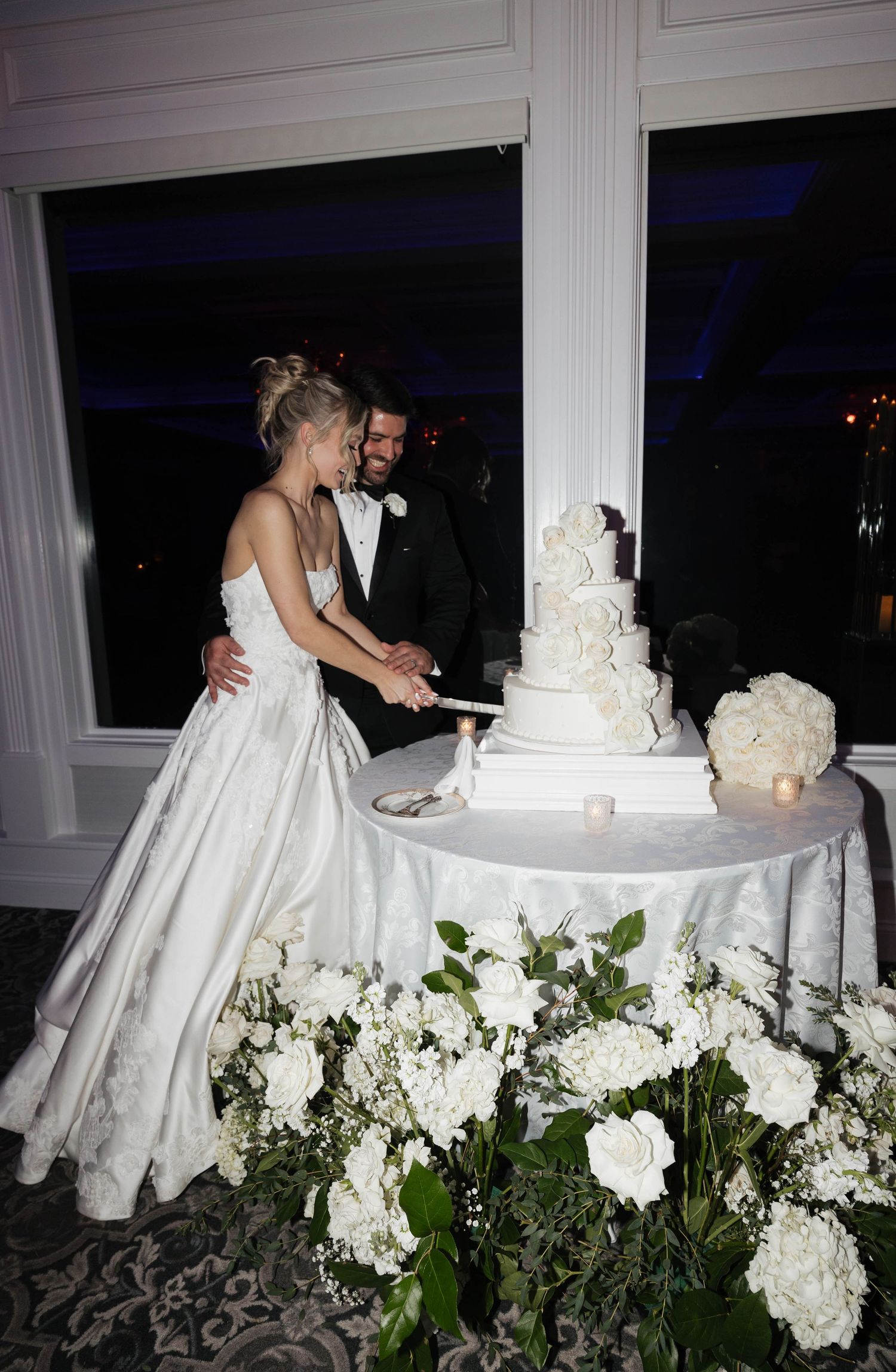 Couple cutting their elegant white wedding cake decorated with flowers at an evening reception.