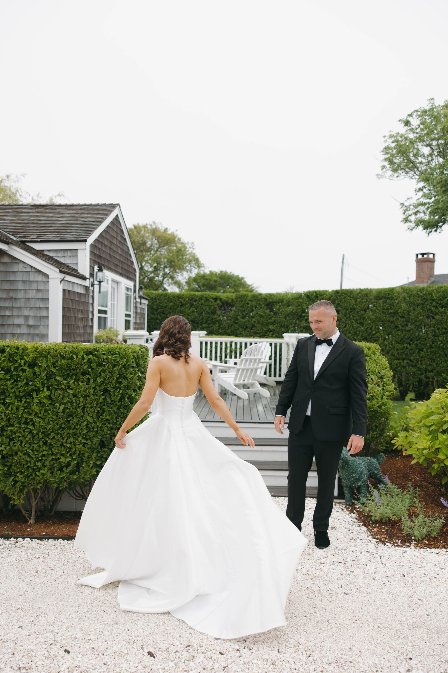 Couple in wedding attire dances and twirls on white gravel garden path.
