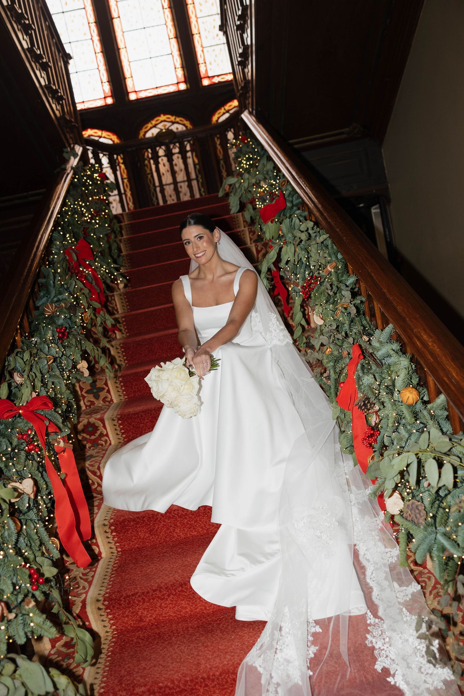 Wedding gown with flowing train photographed on ornate staircase with Christmas decorations and stained glass windows.