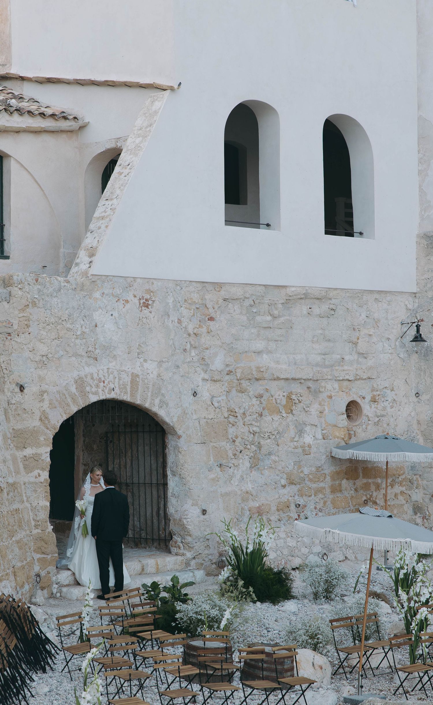 Rustic stone courtyard with folding wooden chairs and white arched architecture.