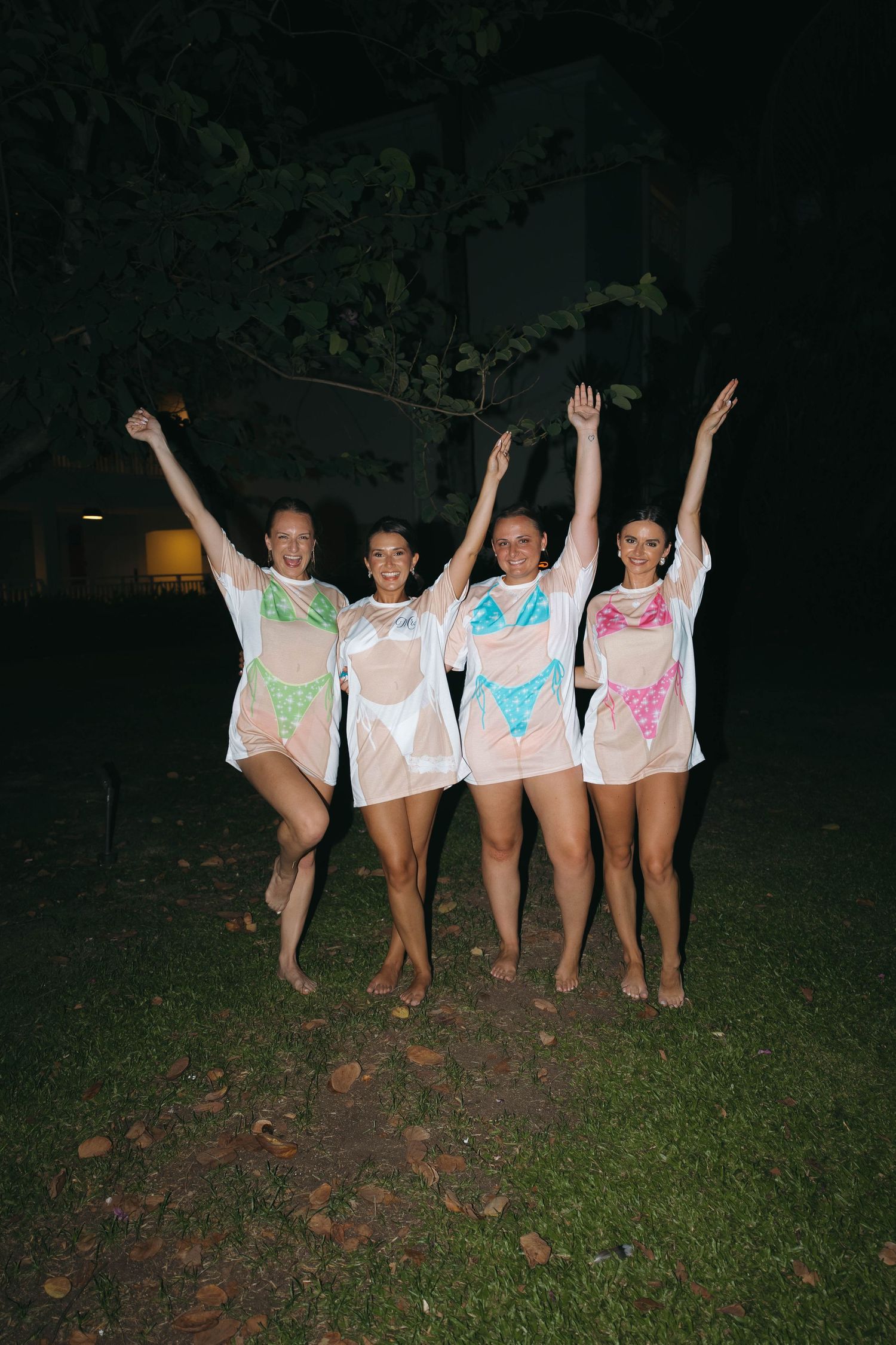 Four women in sheer white cover-ups over colorful swimwear celebrate outdoors at night with arms raised.