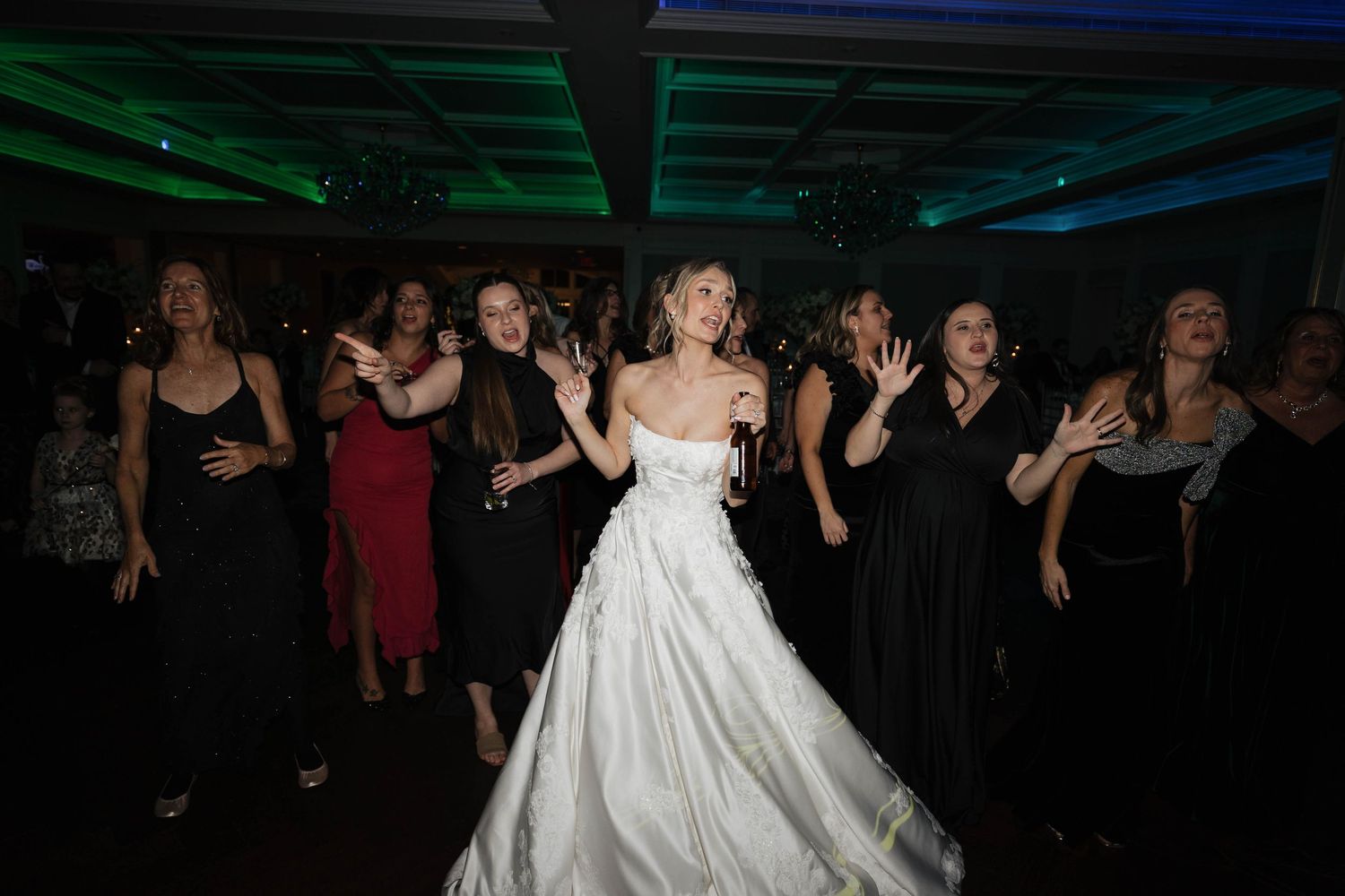 Elegant wedding celebration with guests dancing in formal attire under colored venue lighting.