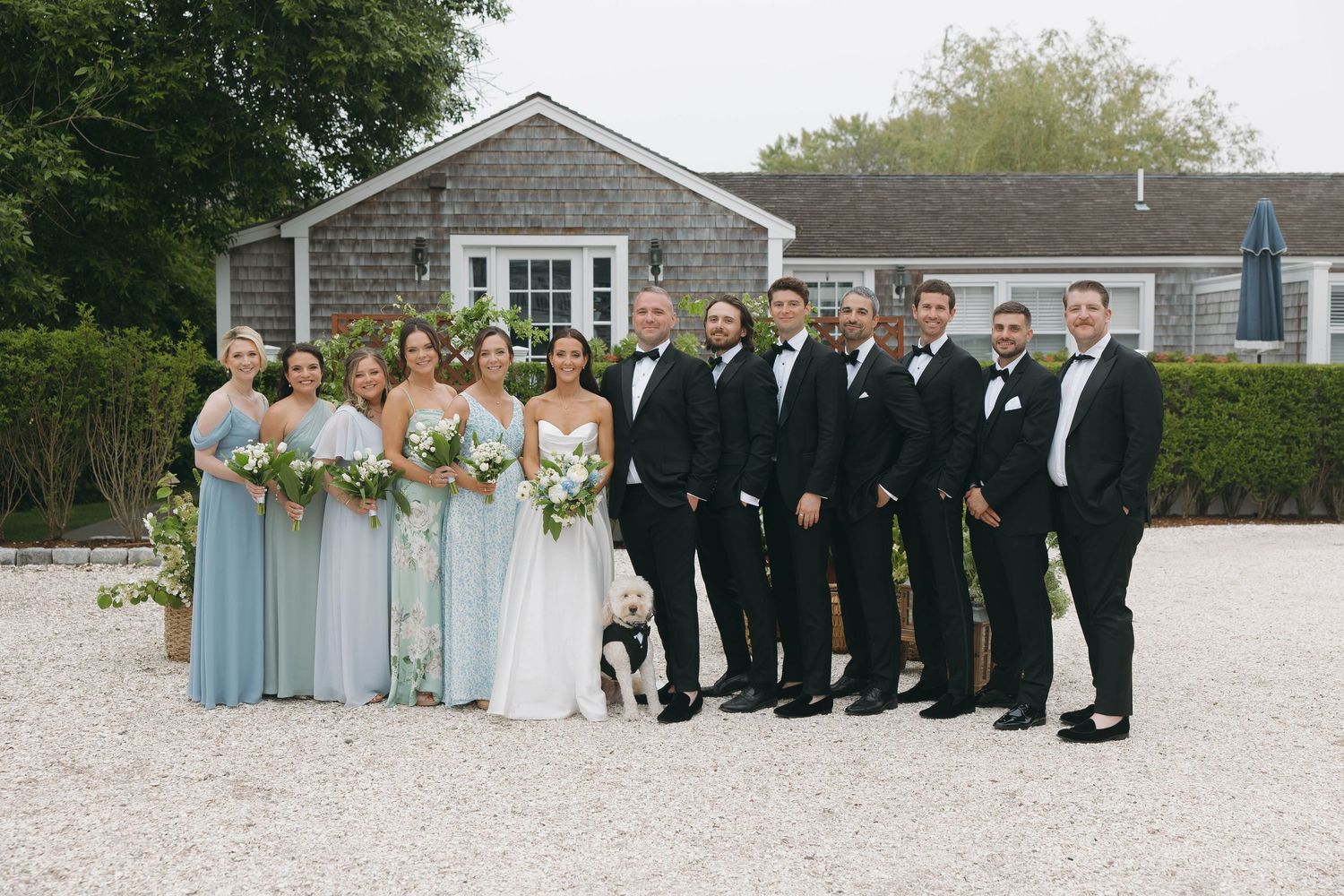 Wedding party poses outdoors on a gravel courtyard wearing light blue bridesmaid dresses and black tuxedos.