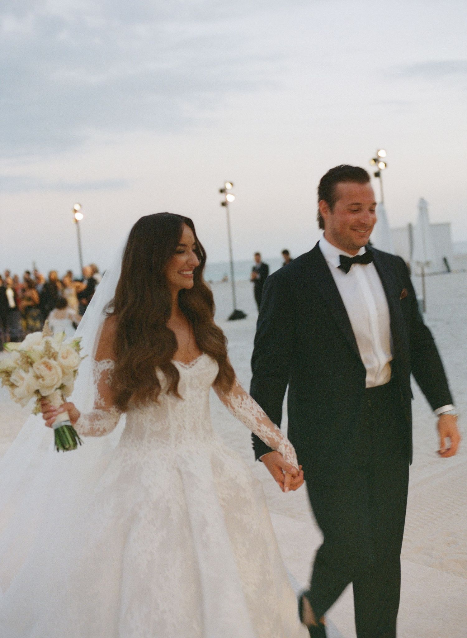 Couple walking along a beach with white floral bouquet against cloudy sky.