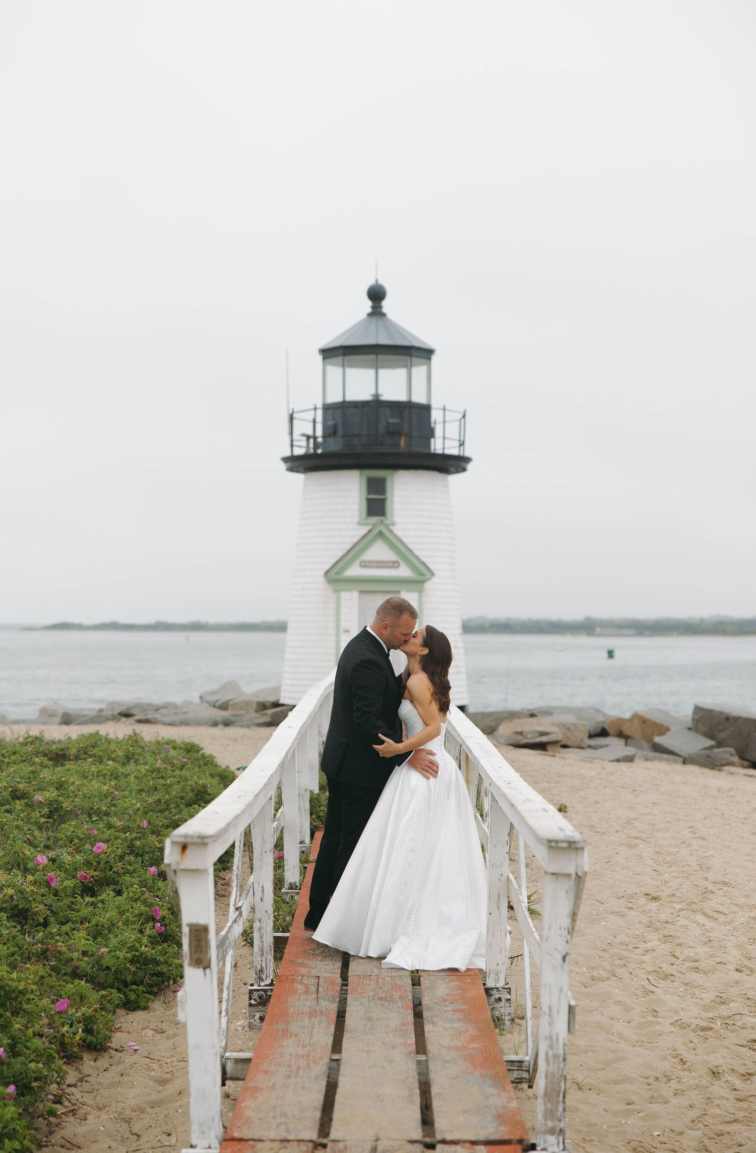Wedding couple shares romantic moment on white lighthouse walkway by the beach.