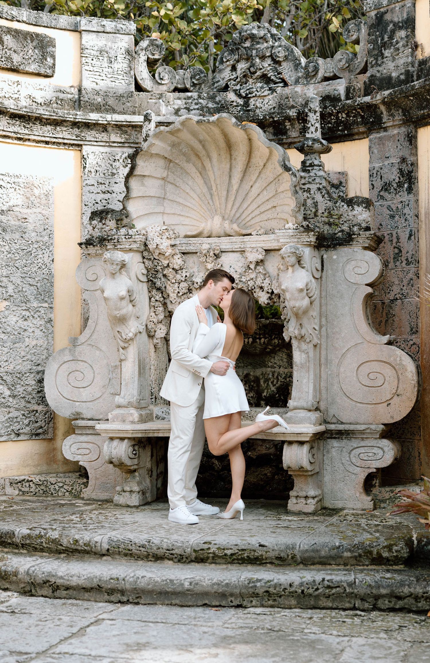 A couple in white attire shares an intimate moment against an ornate stone wall backdrop.
