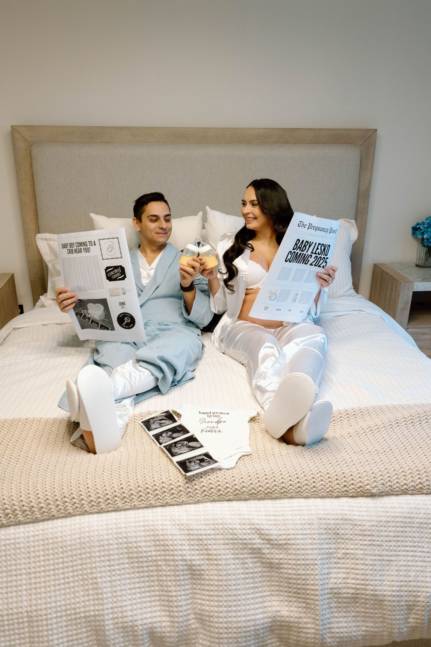 A couple in matching blue and white outfits sits on a bed looking at pregnancy announcements and ultrasound photos.