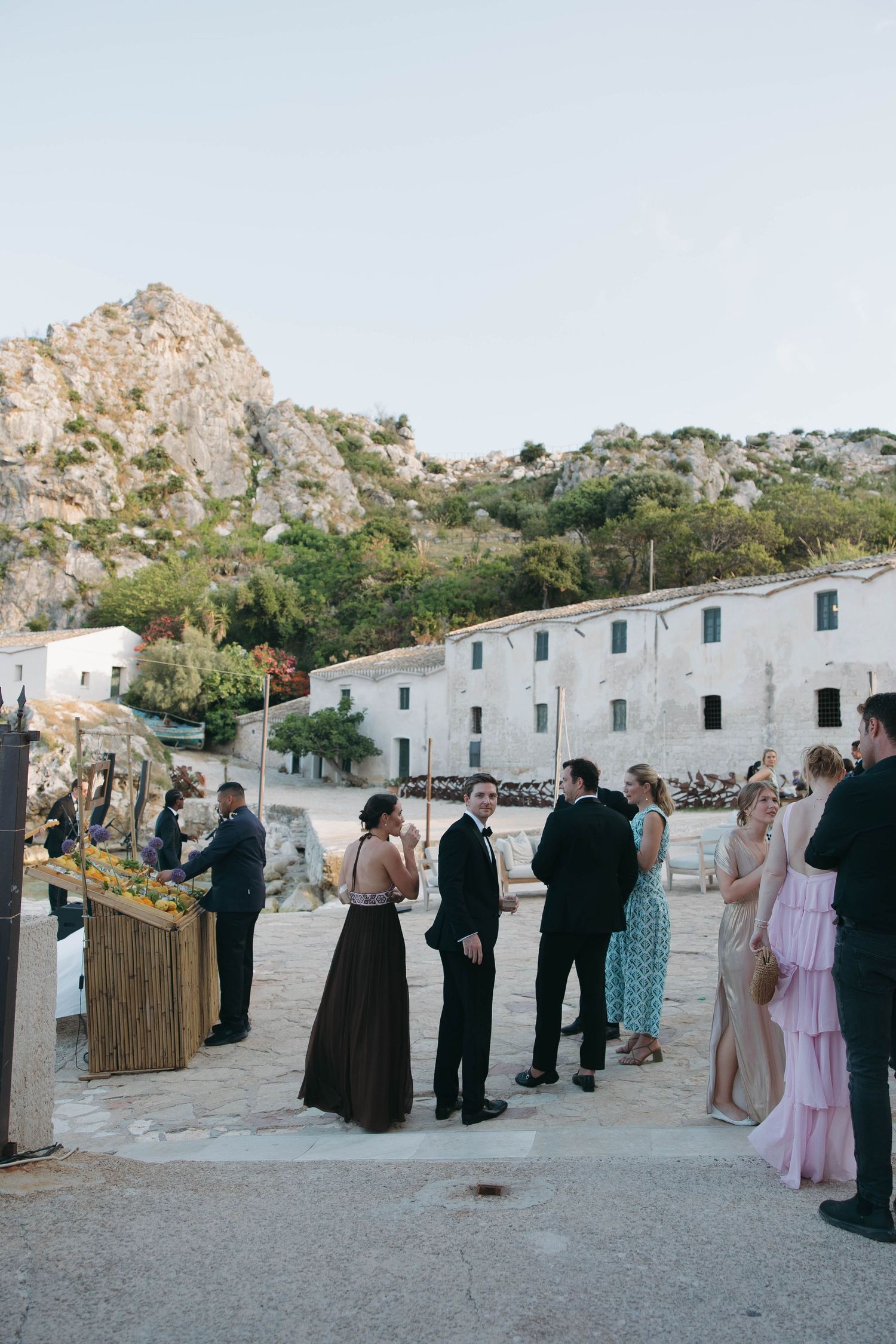 Wedding guests gather outside white buildings against a rocky coastal backdrop.
