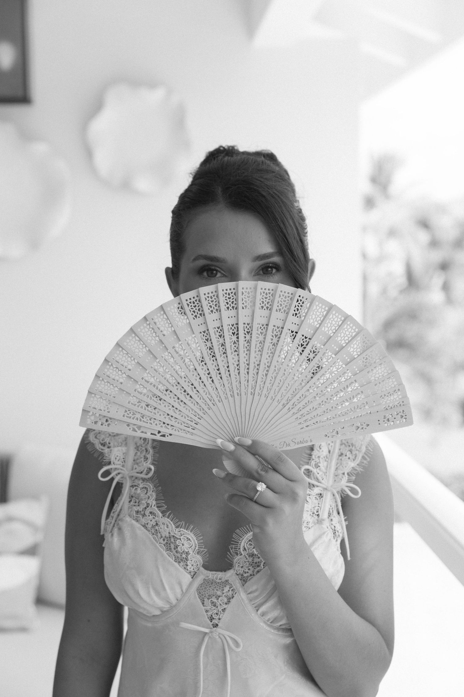 Elegant bride holding a decorative white lace hand fan covering her face in a black and white portrait photo.