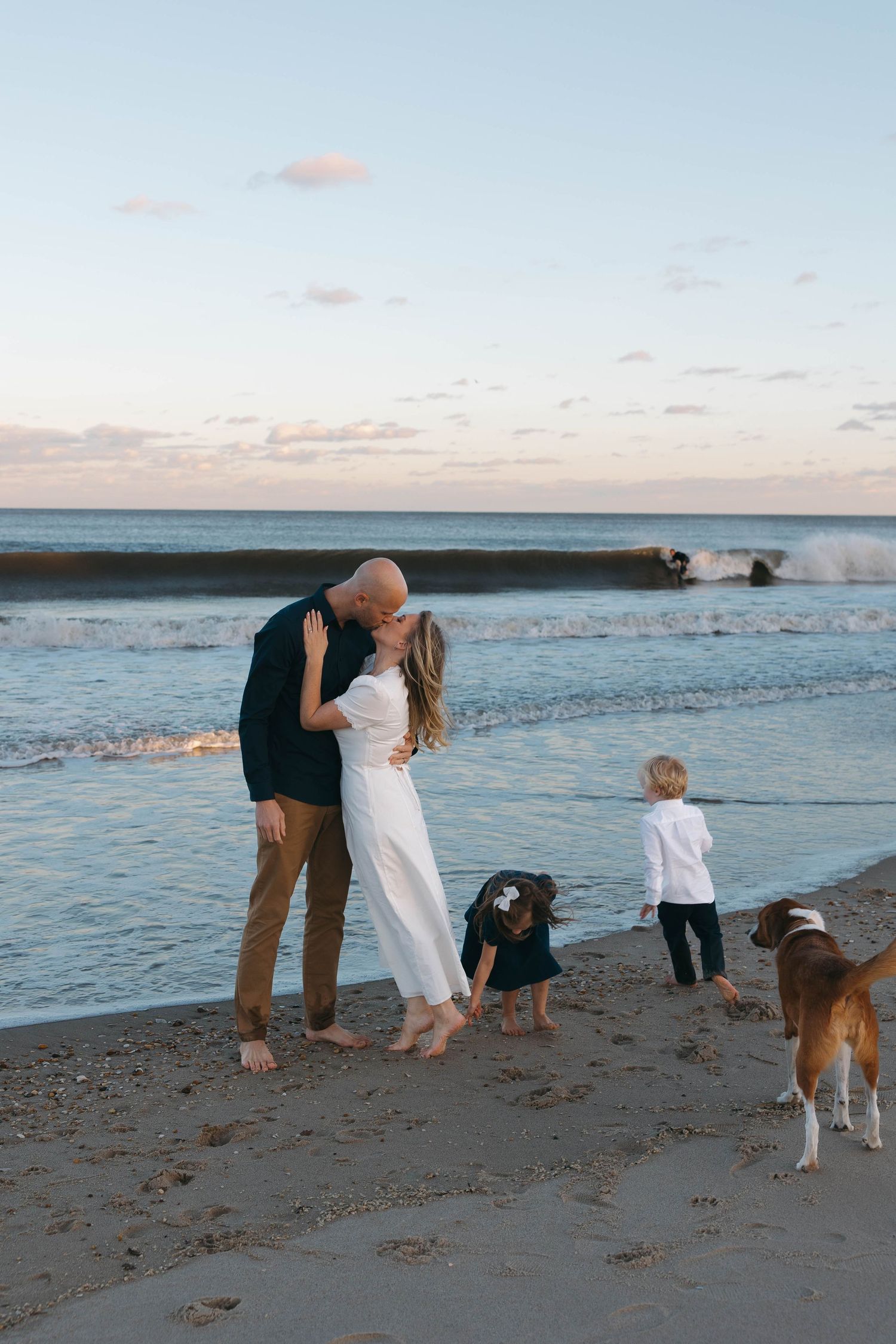 A couple and their dog share a romantic moment on a sandy beach at sunset with gentle waves rolling in.