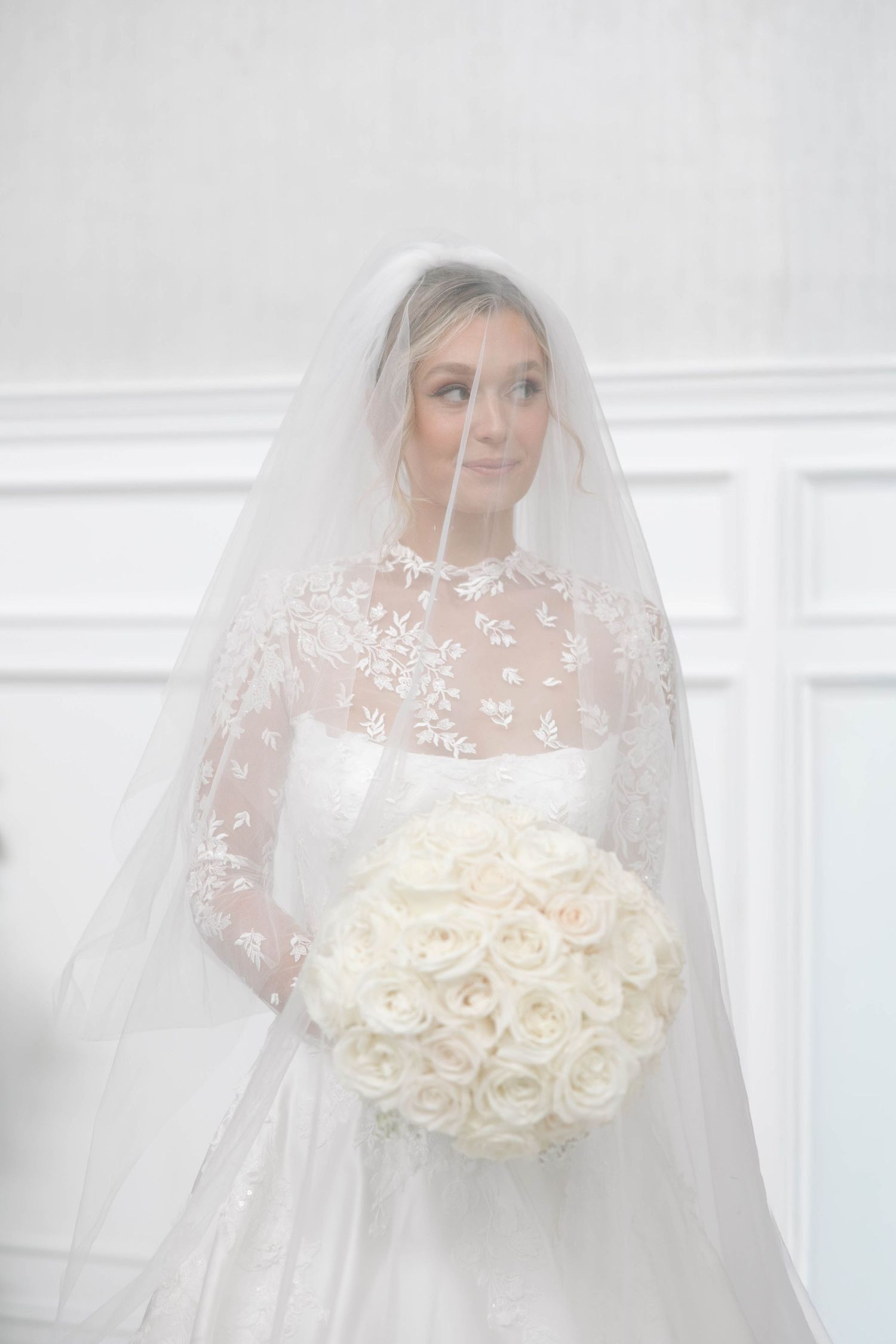 Bride in white gown holds a luxurious bouquet of cream roses against a bright background.