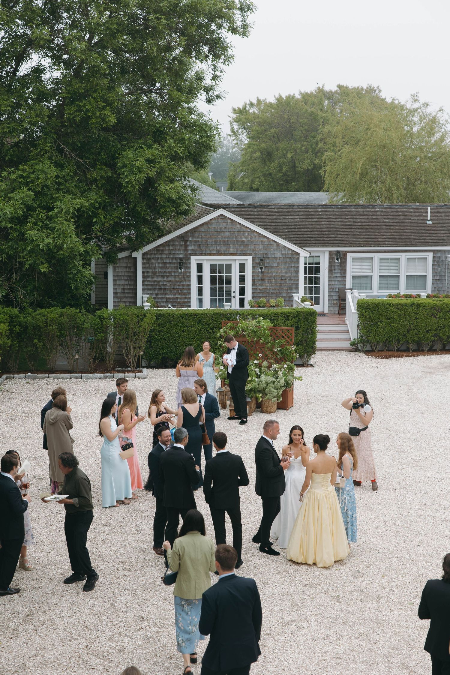 Wedding guests gathered outside a New England style gray shingled building on white gravel.