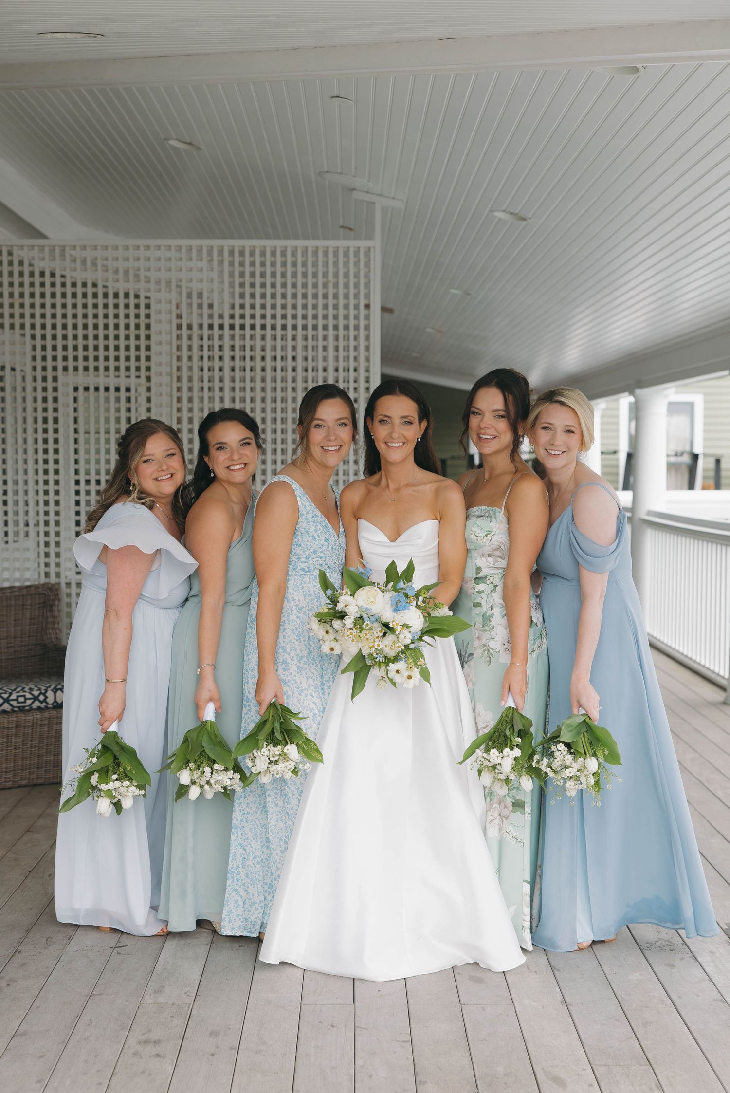 Wedding party in coordinated blue and white dresses gathered for group photos on bright porch.