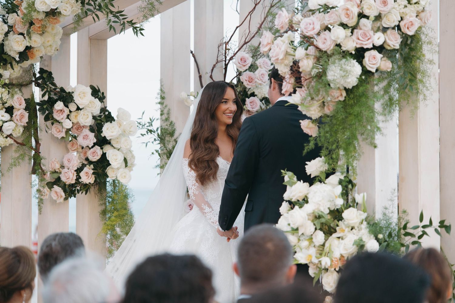 Wedding guests gather beneath an elegant floral-adorned white pergola during a ceremony.