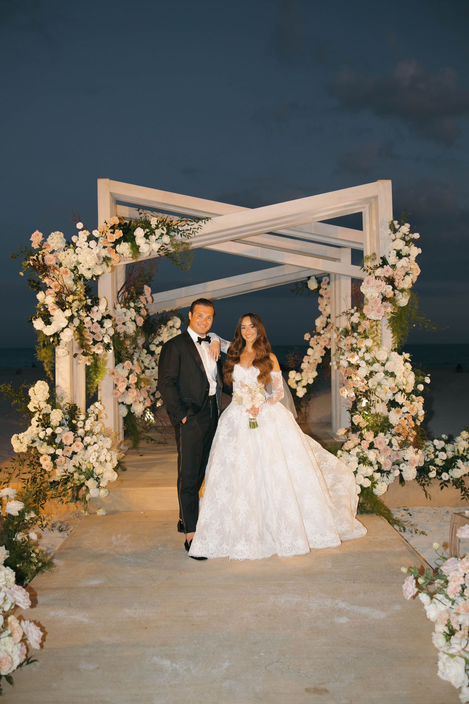 Romantic beach wedding ceremony under a white pergola adorned with lush floral arrangements.