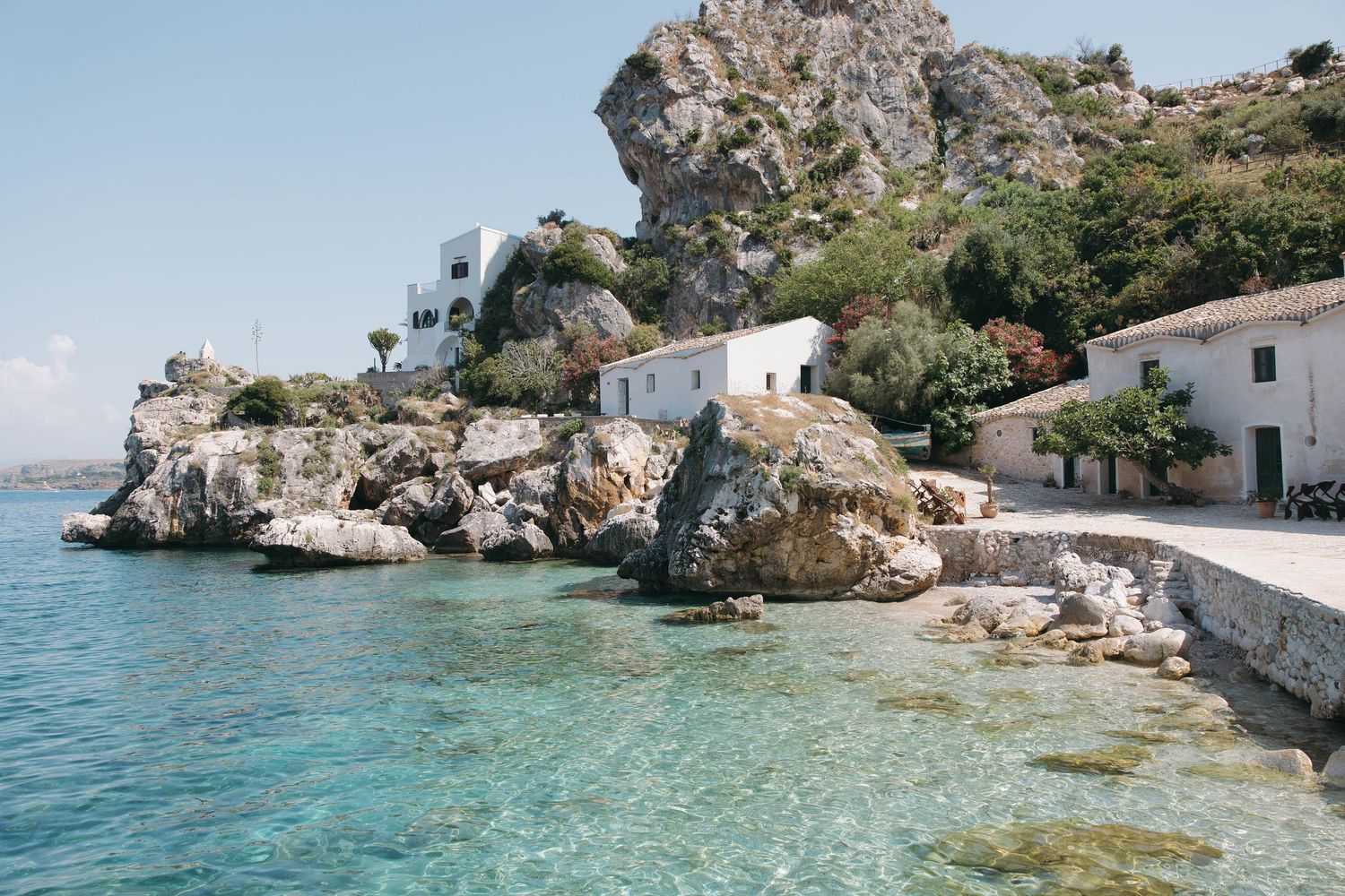 Rocky coastline with crystal clear turquoise waters and white buildings along the Mediterranean Sea shore.