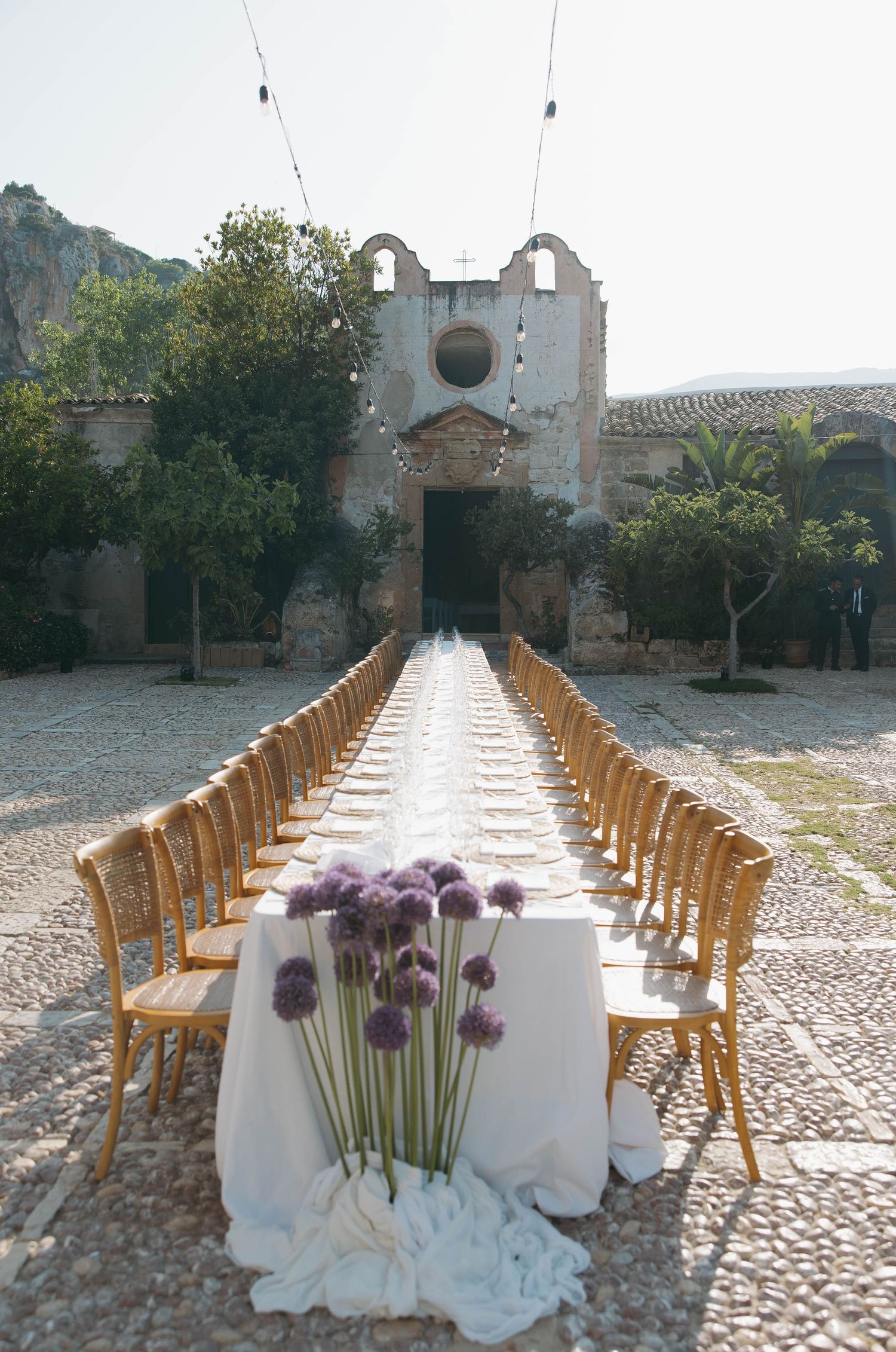 Long white dining table with wooden chairs set up outdoors with purple allium flower centerpieces.