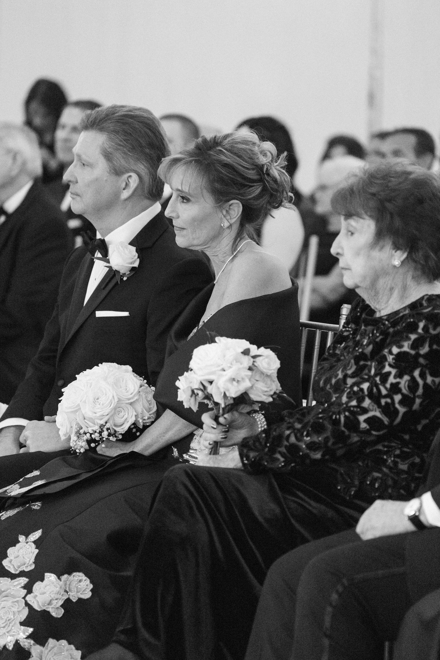 Guests in formal black attire seated with white floral arrangements at elegant ceremony.