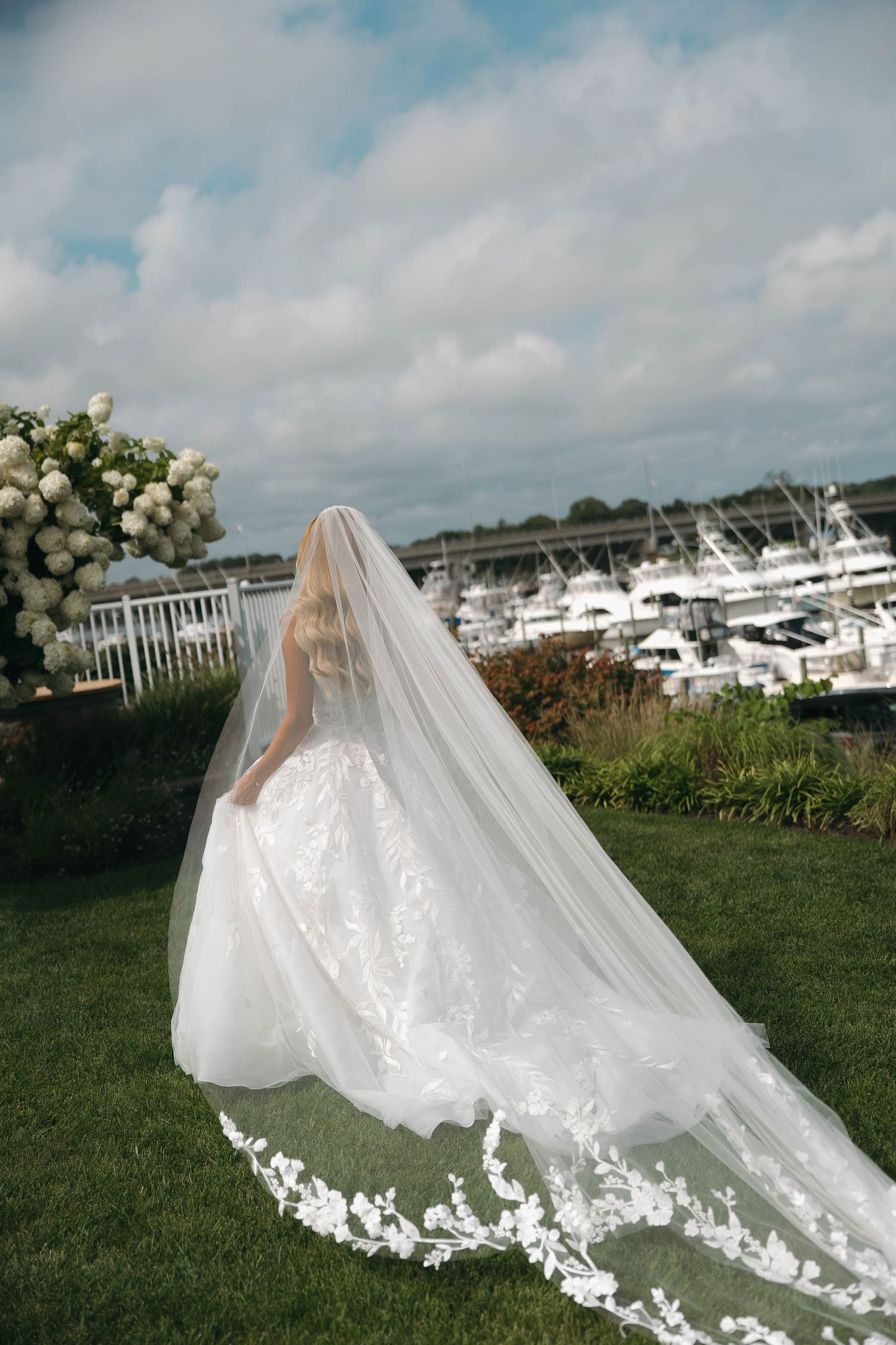 Bride in a stunning lace ball gown with a long floral-trimmed cathedral veil, standing by a marina on a cloudy day.