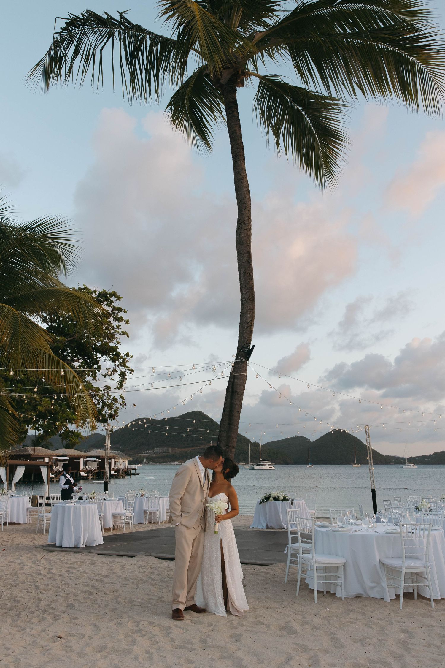 Couple kissing under a palm tree on a beach wedding venue at sunset with white reception tables.