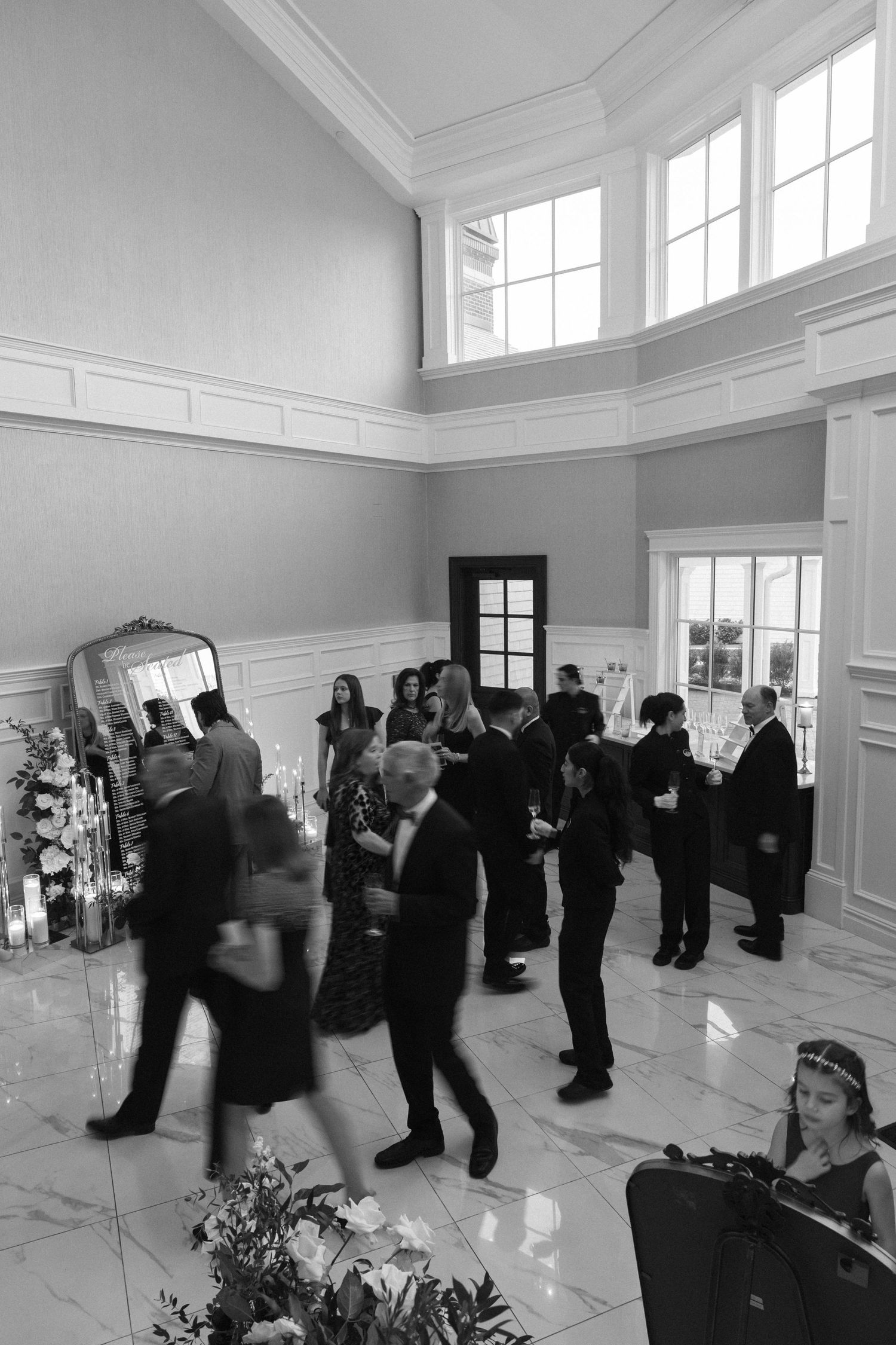 A group of people gathered in a bright white room with large windows for what appears to be a social event.