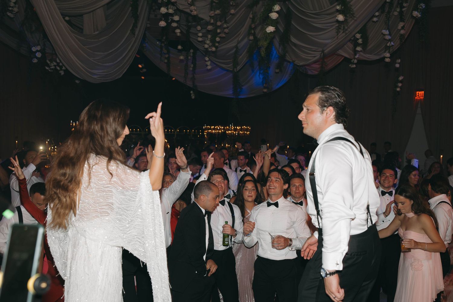 Large group of wedding guests dancing together under dramatic lighting at an evening reception.