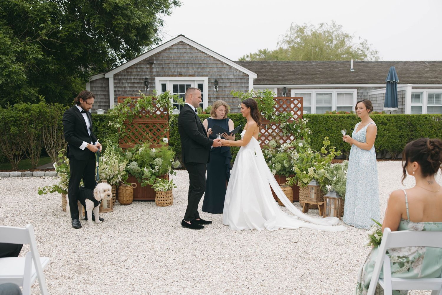 Outdoor wedding ceremony in progress at a coastal venue with white chairs and potted plants.