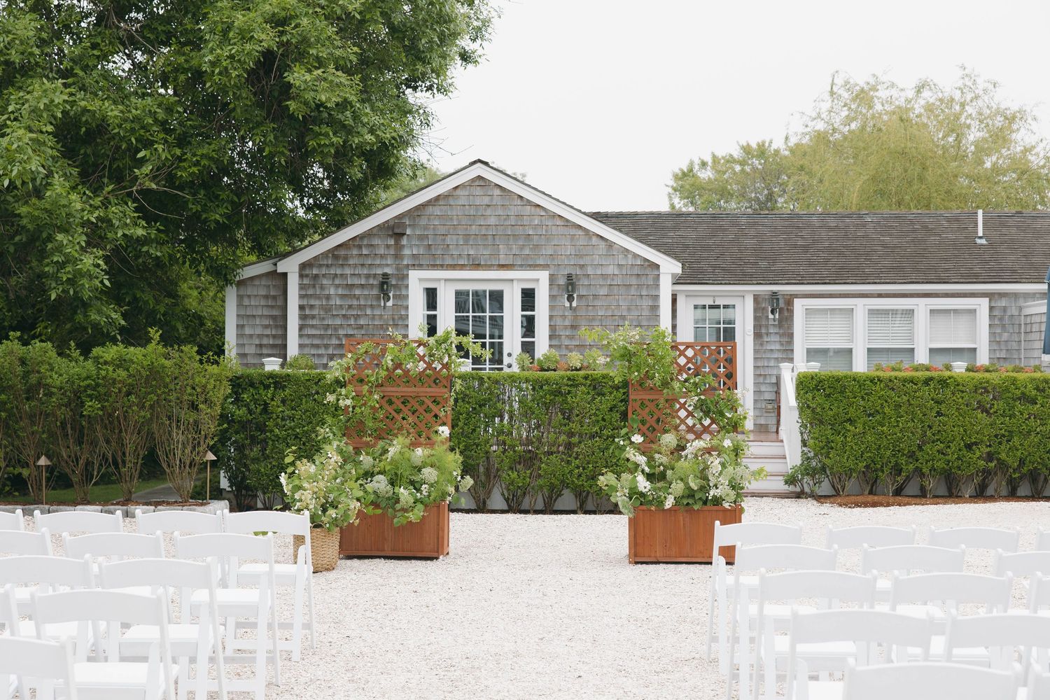 Outdoor wedding venue with white chairs set up on a gravel surface in front of gray shingled beach houses.