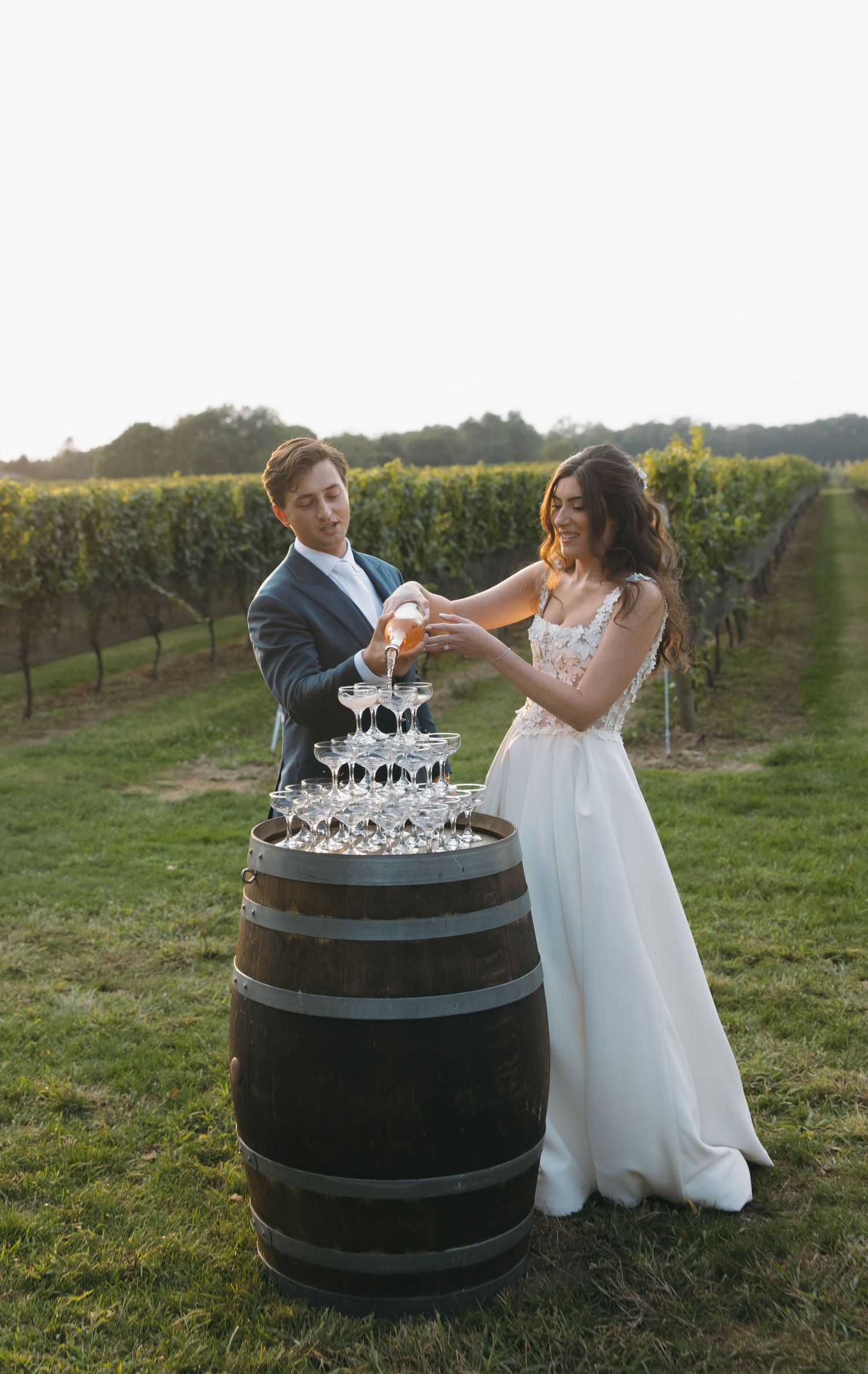 A couple playfully interacts around a wine barrel in a scenic vineyard setting.