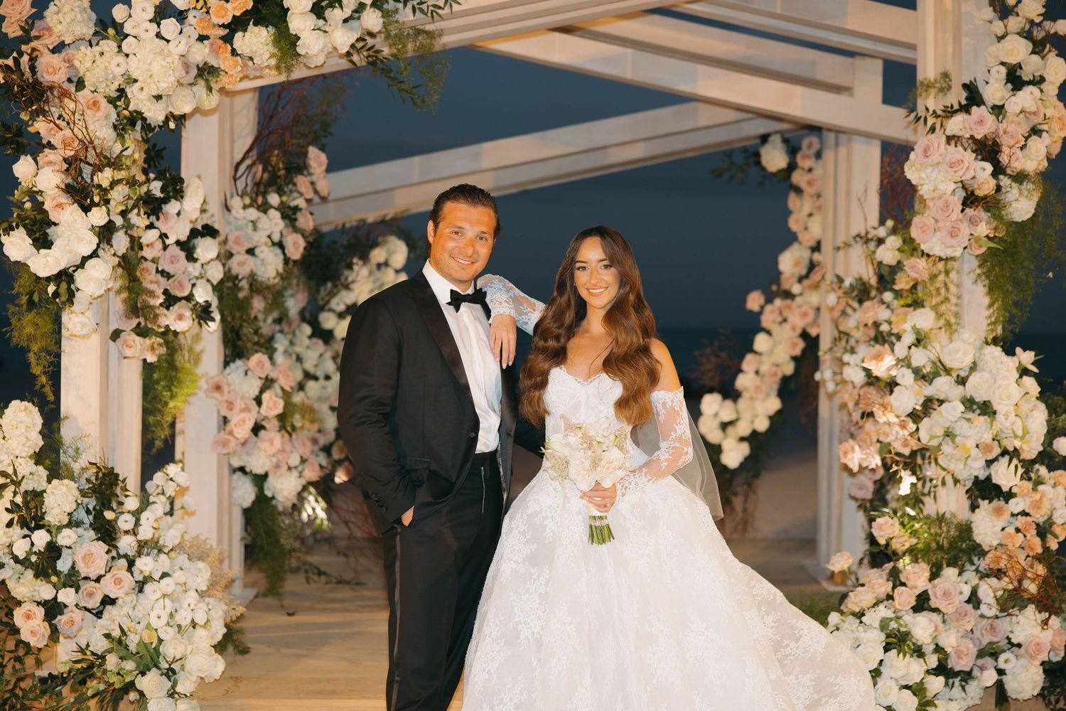 White floral arrangements decorate a beachfront wedding ceremony structure at sunset.