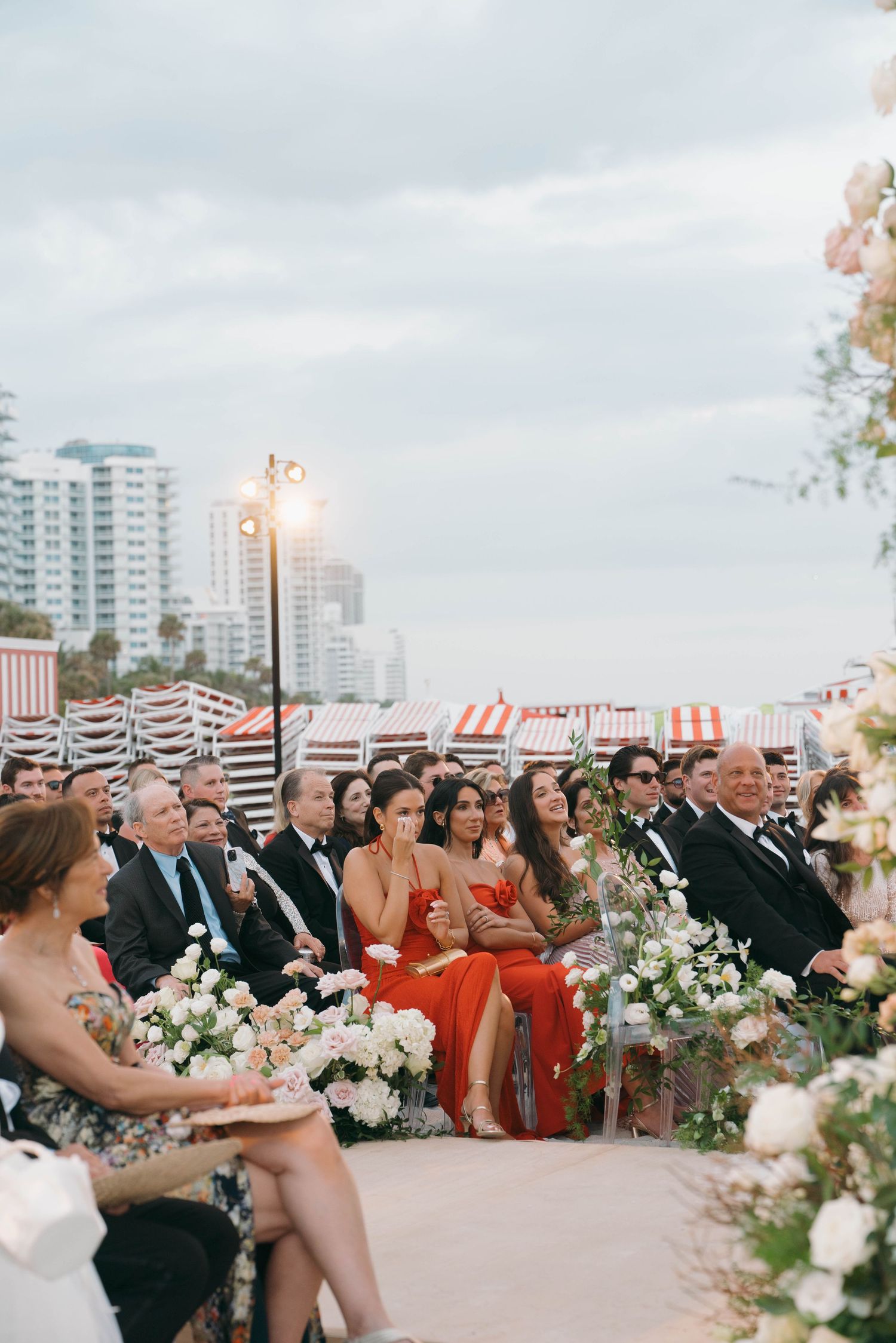 Wedding guests seated outdoors with city buildings visible in the background and floral decorations.
