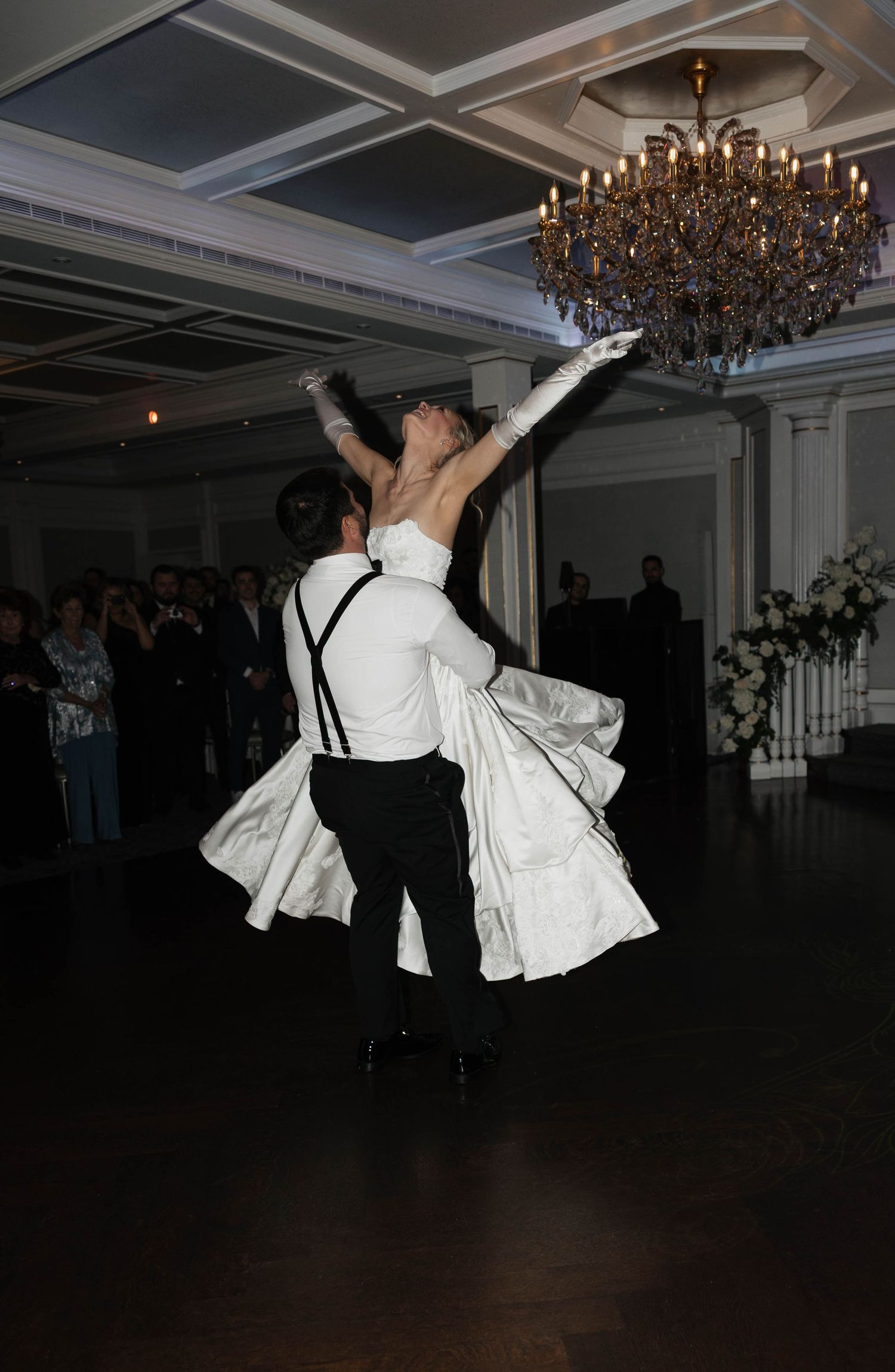 Wedding couple executes a dramatic dance lift in a dimly lit ballroom with chandeliers overhead.
